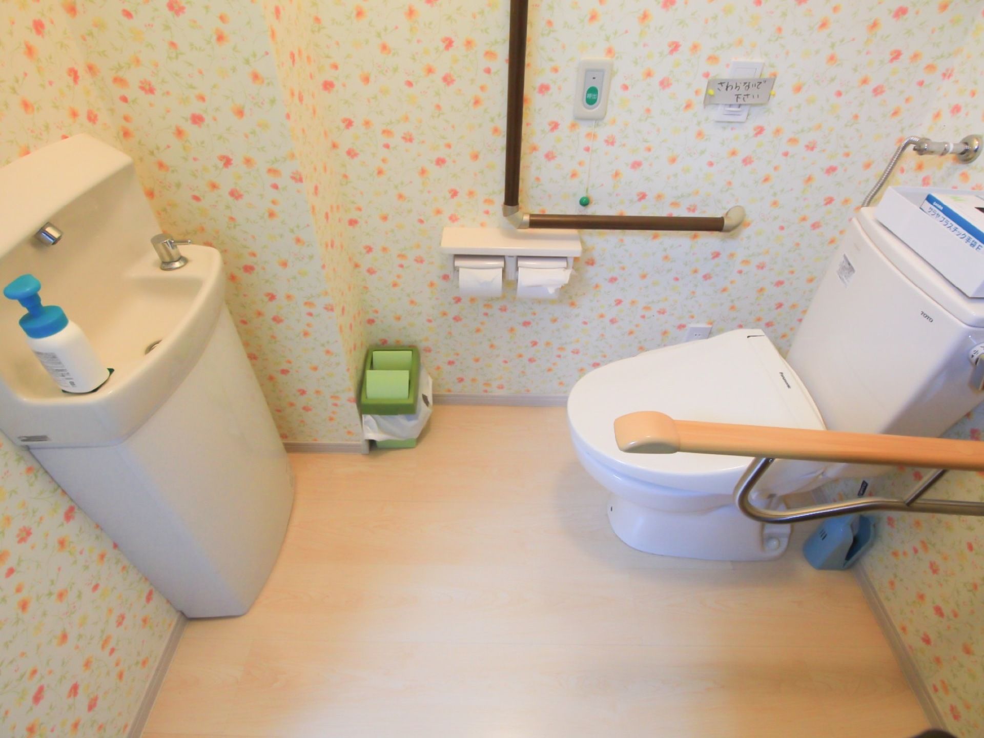 可愛らしい壁紙のトイレである。L型や可動式手すり、洗面台が完備されており、呼び出しボタンが壁に設置されている。