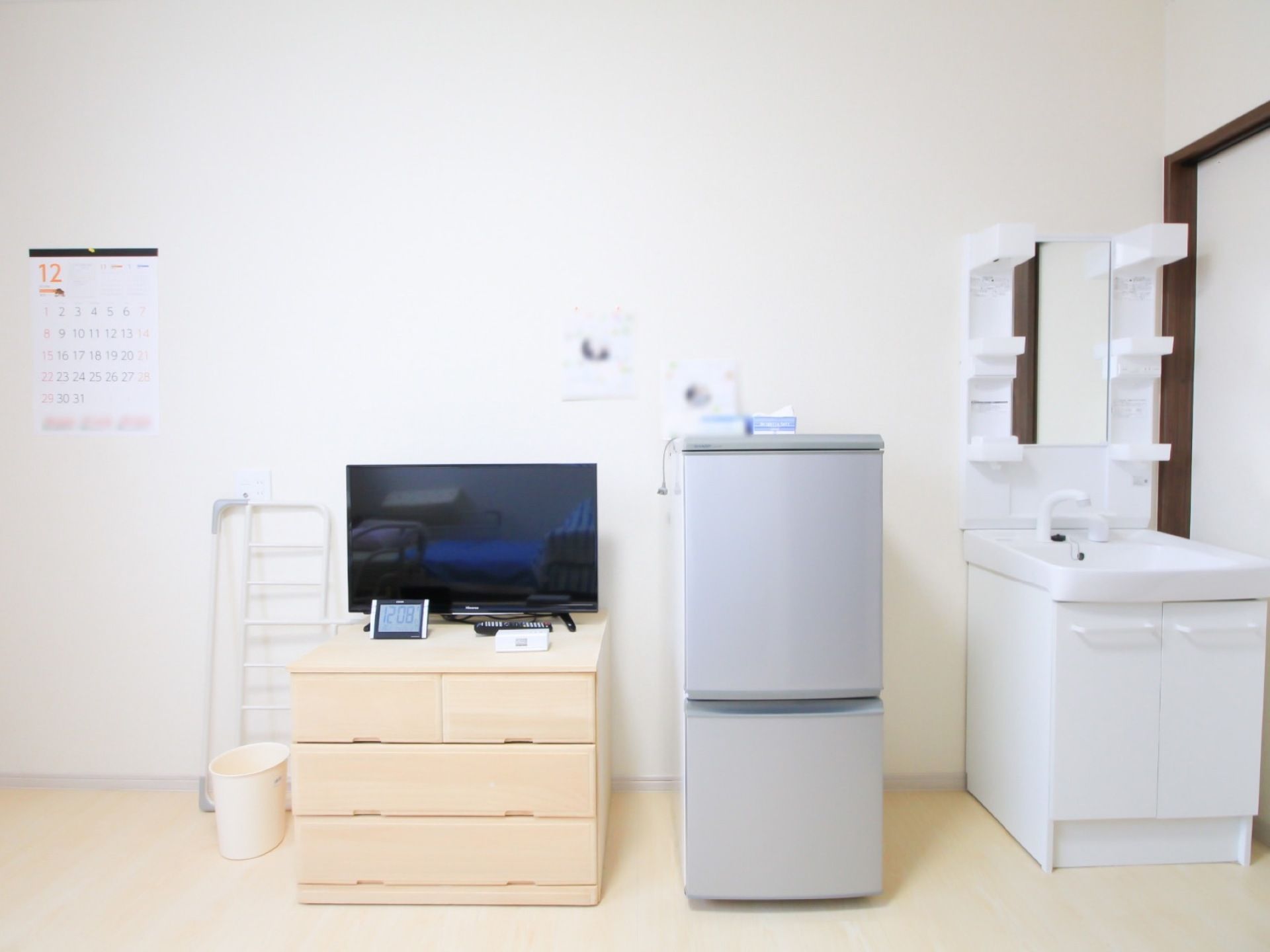 洗面化粧台が完備された居室である。壁際に冷蔵庫や収納家具、テレビが設置されており、ゴミ箱が置かれている。