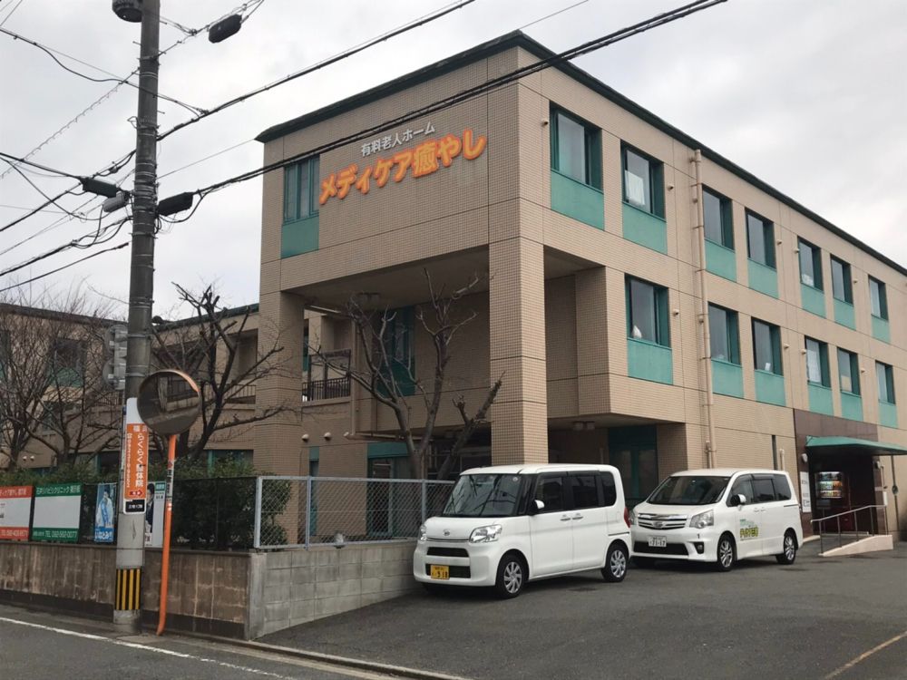 3階建ての建物の玄関には屋根があり、手すりも付いている。敷地内には駐車場があり、車が数台止まっている。