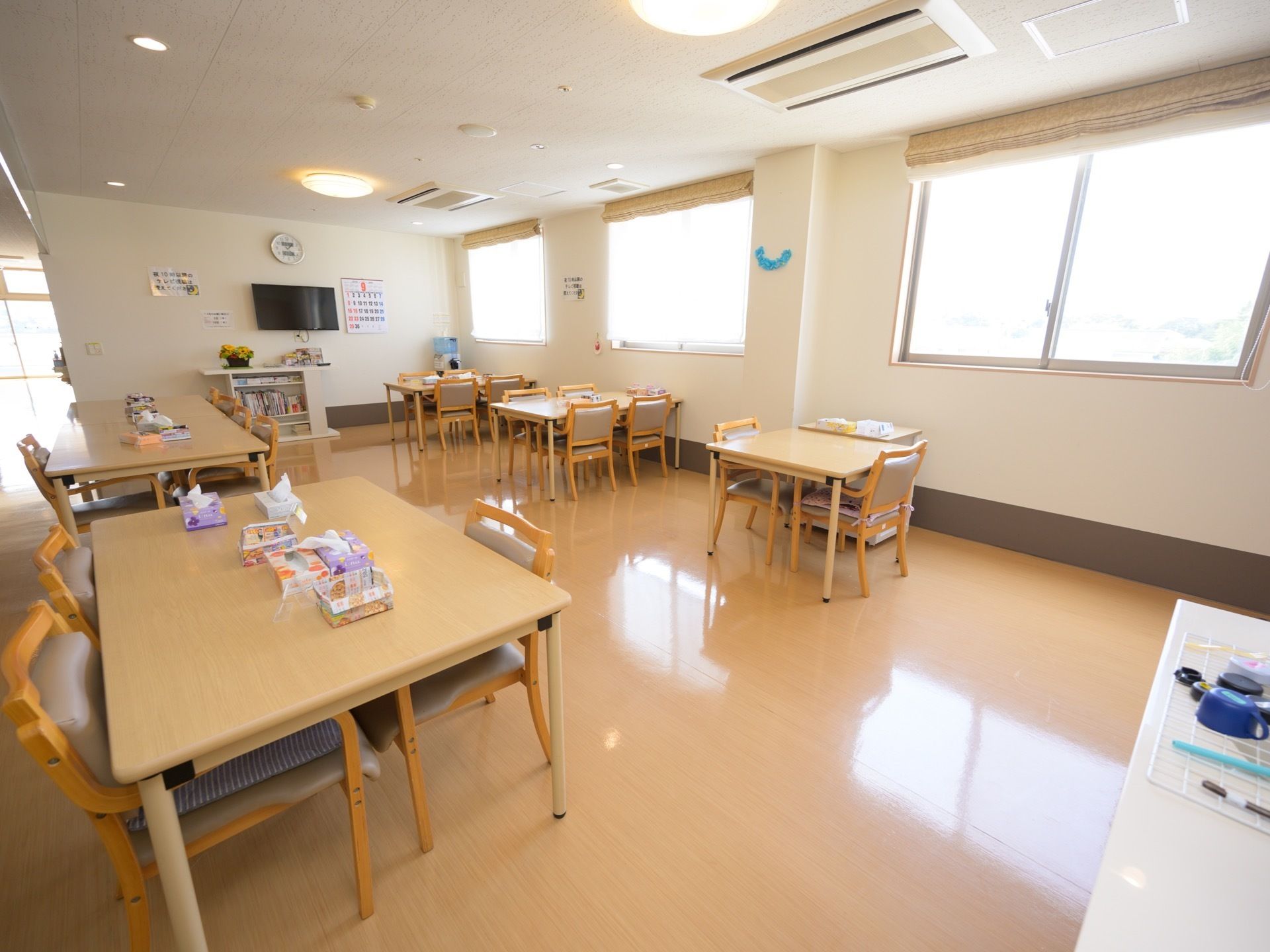 窓から日差しが差し込む明るく開放的な食堂である。テーブルと椅子が用意されており、壁にテレビがある。