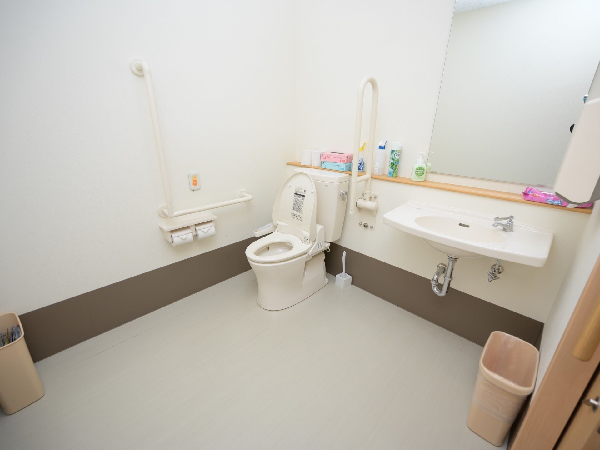 広いスペースが確保された共用トイレである。洗面台や手すり、呼び出しボタンが完備されている。ゴミ箱が出入り口近くに置かれている。