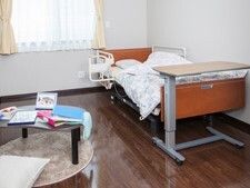 レースカーテンとカーテンが付いた窓があるフローリングの部屋には介護ベッドやテーブルがある。