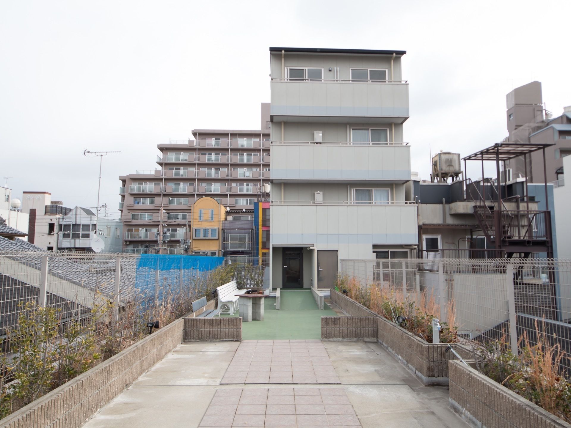 隣の家の屋根くらいの高さにあるアプローチの先に玄関がある。アプローチの両側にはフェンスが設けられている。