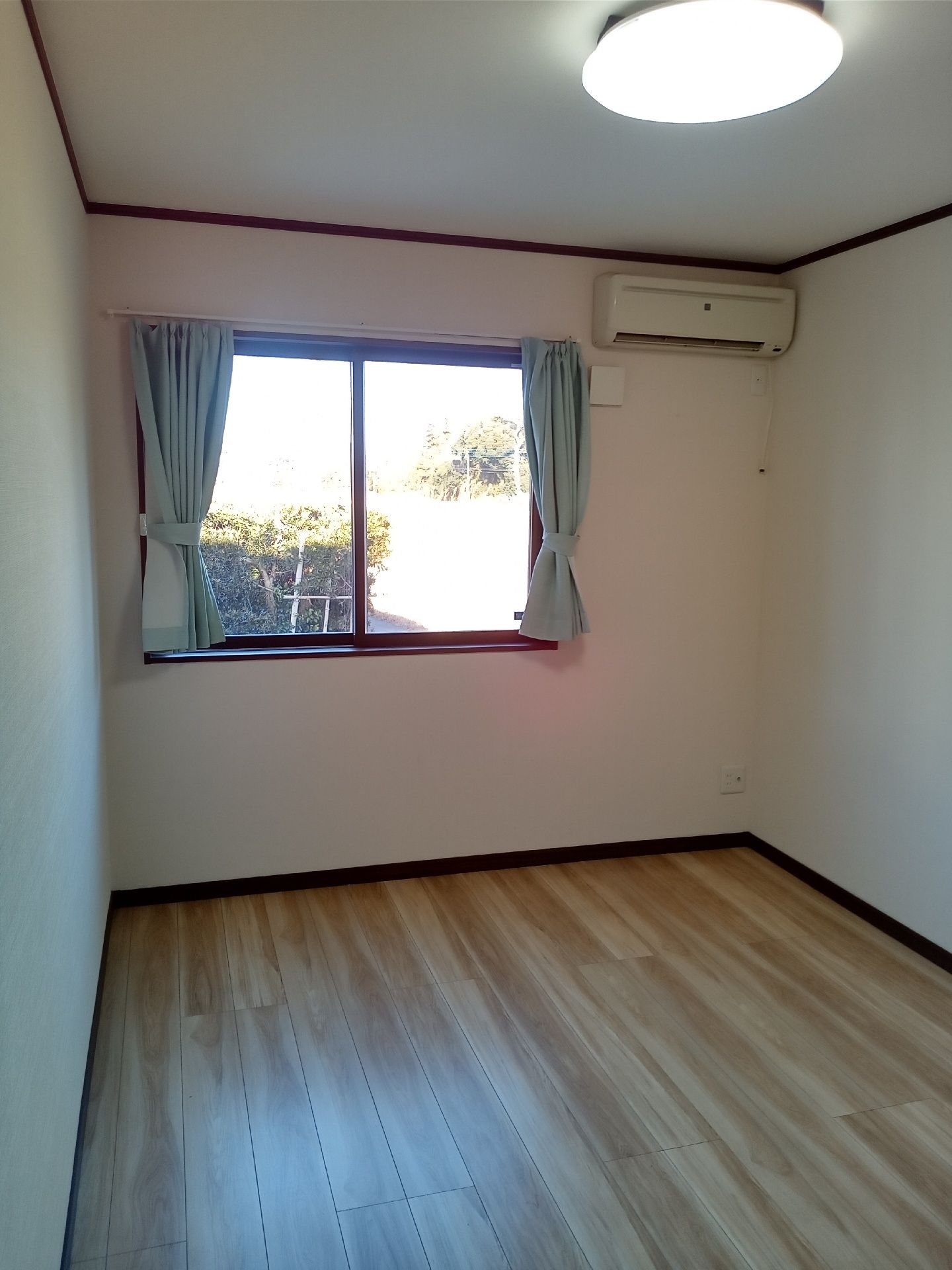 カーテンが設置された大きな窓が付いたフローリングの部屋には家具は置かれていない。照明も付いている。