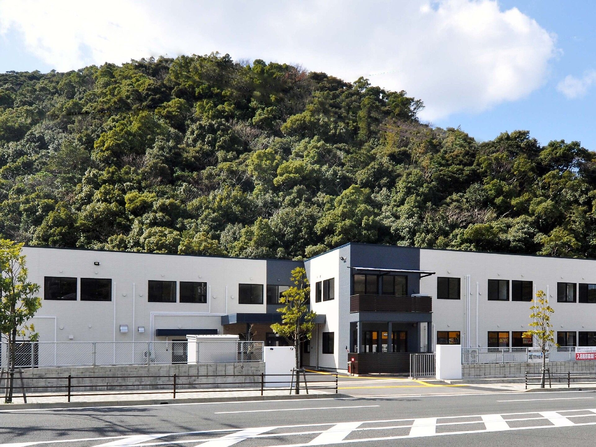 緑に囲まれた2階建ての建物。敷地内には駐車場があり、玄関部分には屋根がある。