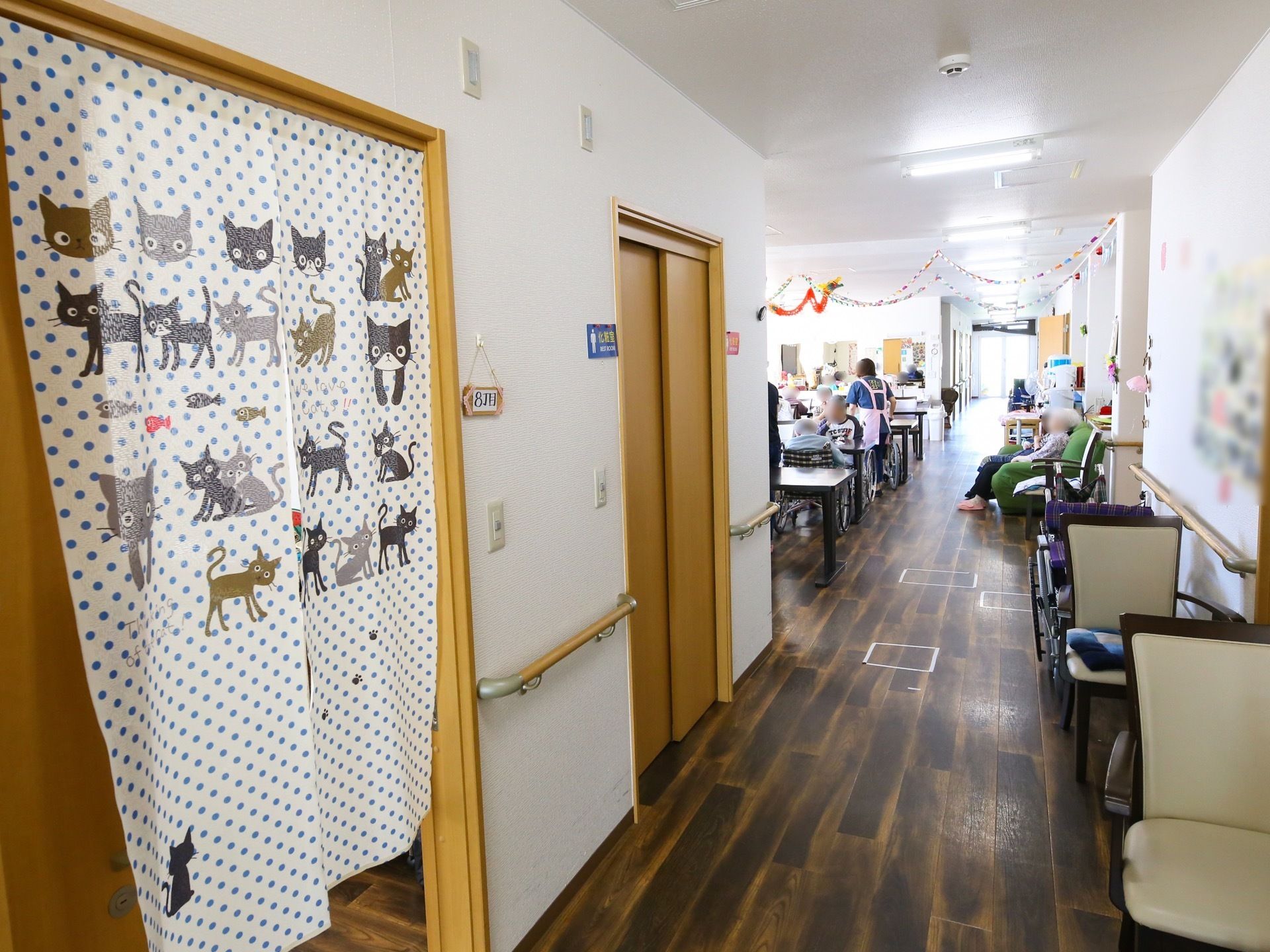 食堂に続いているフローリングの廊下がある。入居者様が壁際に置かれたソファや、ダイニングテーブルについてくつろいでいる。