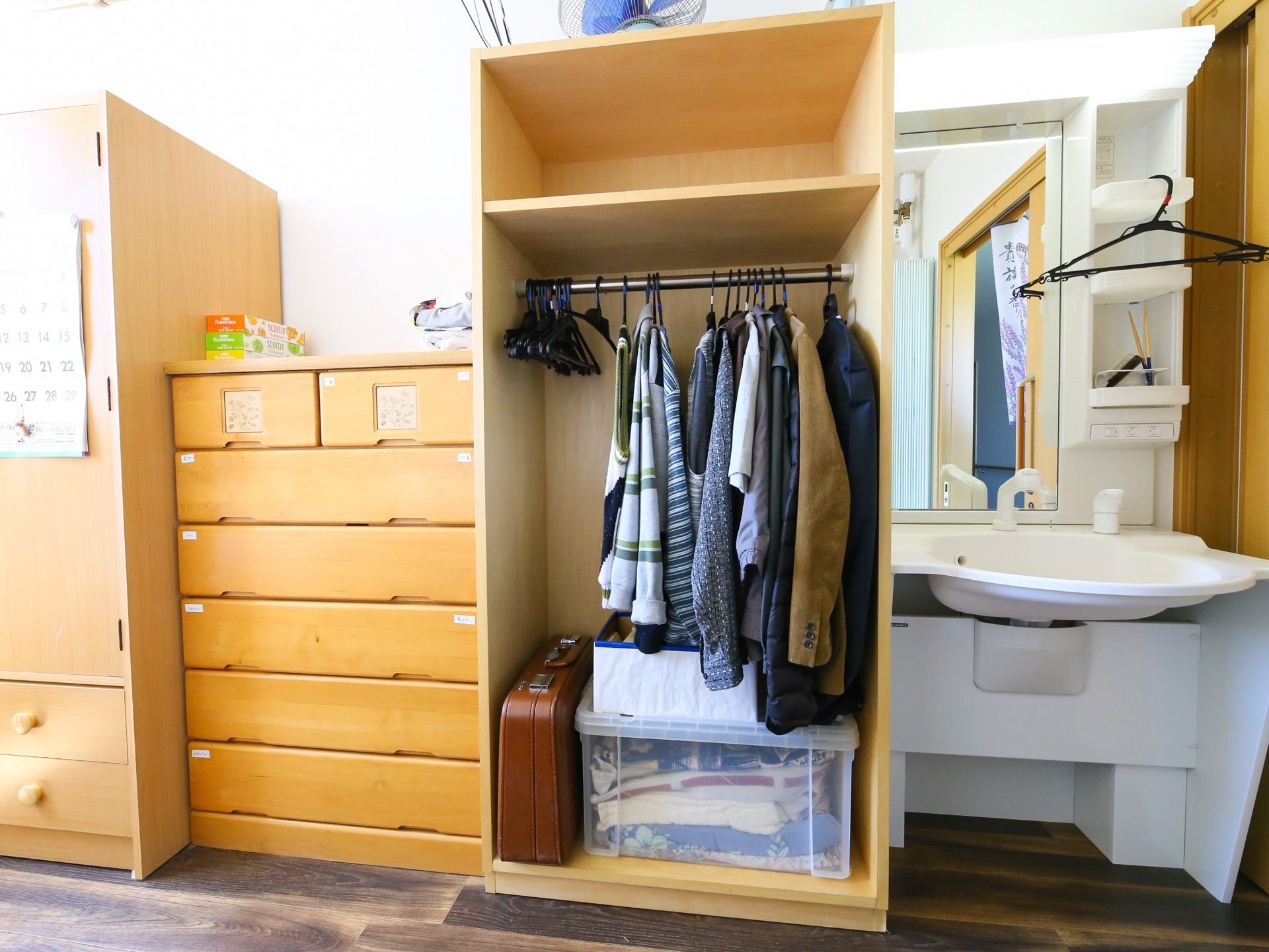フローリングの部屋に洗面台やタンスが並んでいる。クローゼットには上着が掛けられているほか、カバンや衣装ケースも入っている。