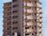 ブラウン系の鉄骨造りの建物は縦に長い大きなマンションタイプの外観で、建物の1階部分が広くなっていて、周りには木が植えられている。