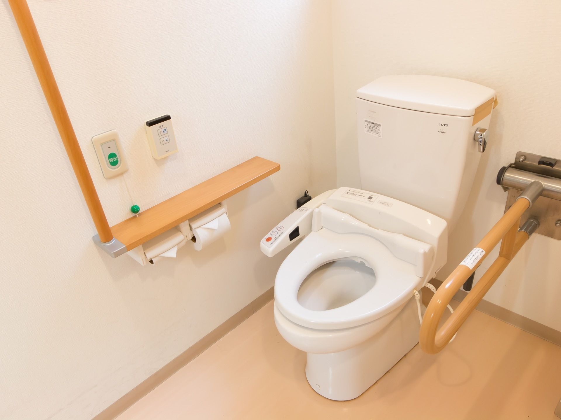 フローリングの床に白い壁のトイレである。便座横に温水洗浄便座の操作パネルがある。壁に非常ボタンがある。
