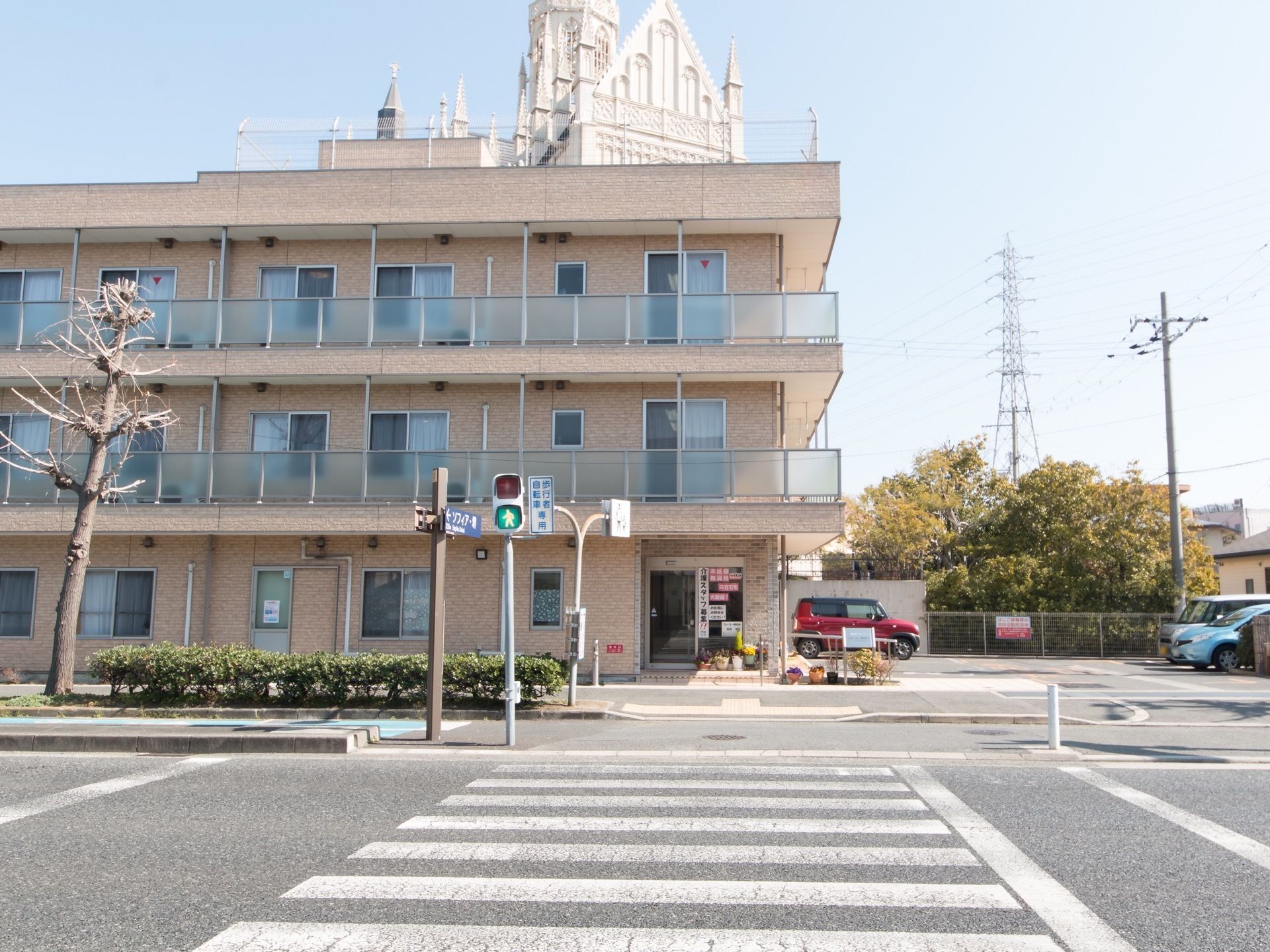 薄いベージュの建物で３階建てである。施設の横に駐車場があり、車が停まっている。横断歩道があり、青になっている。