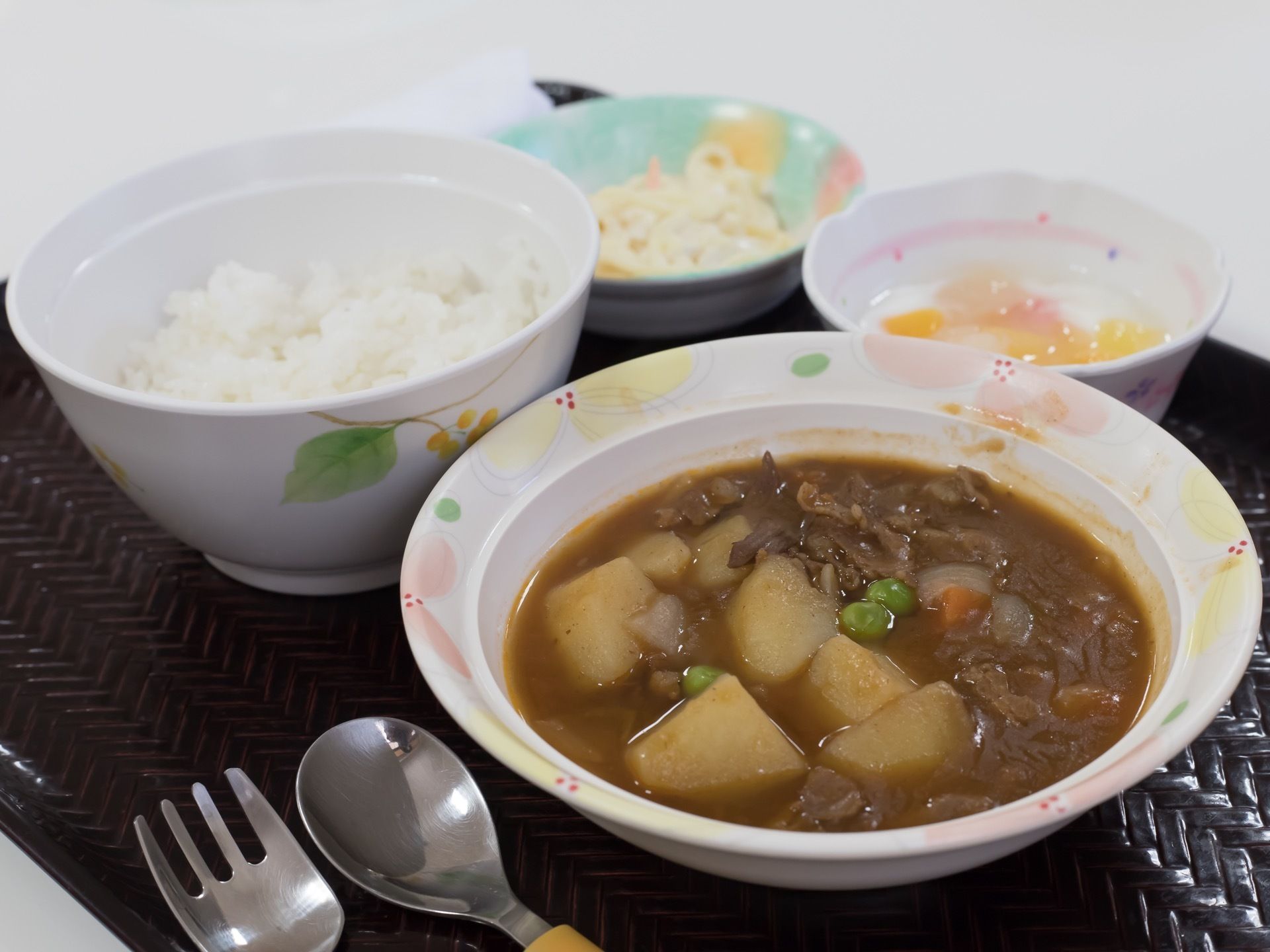 黒いトレーに食事が配膳されている。メインはカレーで、その横にご飯がある。奥に小鉢が２つある。スプーンとフォークがある。
