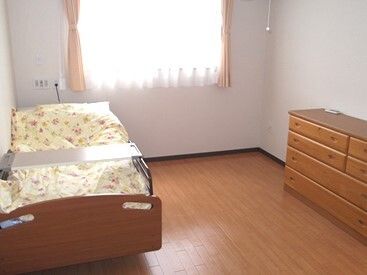 花柄の寝具がセットされた介護ベッドが置かれたフローリングの居室。大きな窓にはカーテンが付いていて、チェストも置かれている。