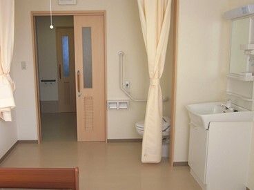 緑の呼び出しボタンや手すりが設置されたトイレ。カーテンで開け閉めするタイプで、居室のドアはスライド式。洗面所やベッドもある。
