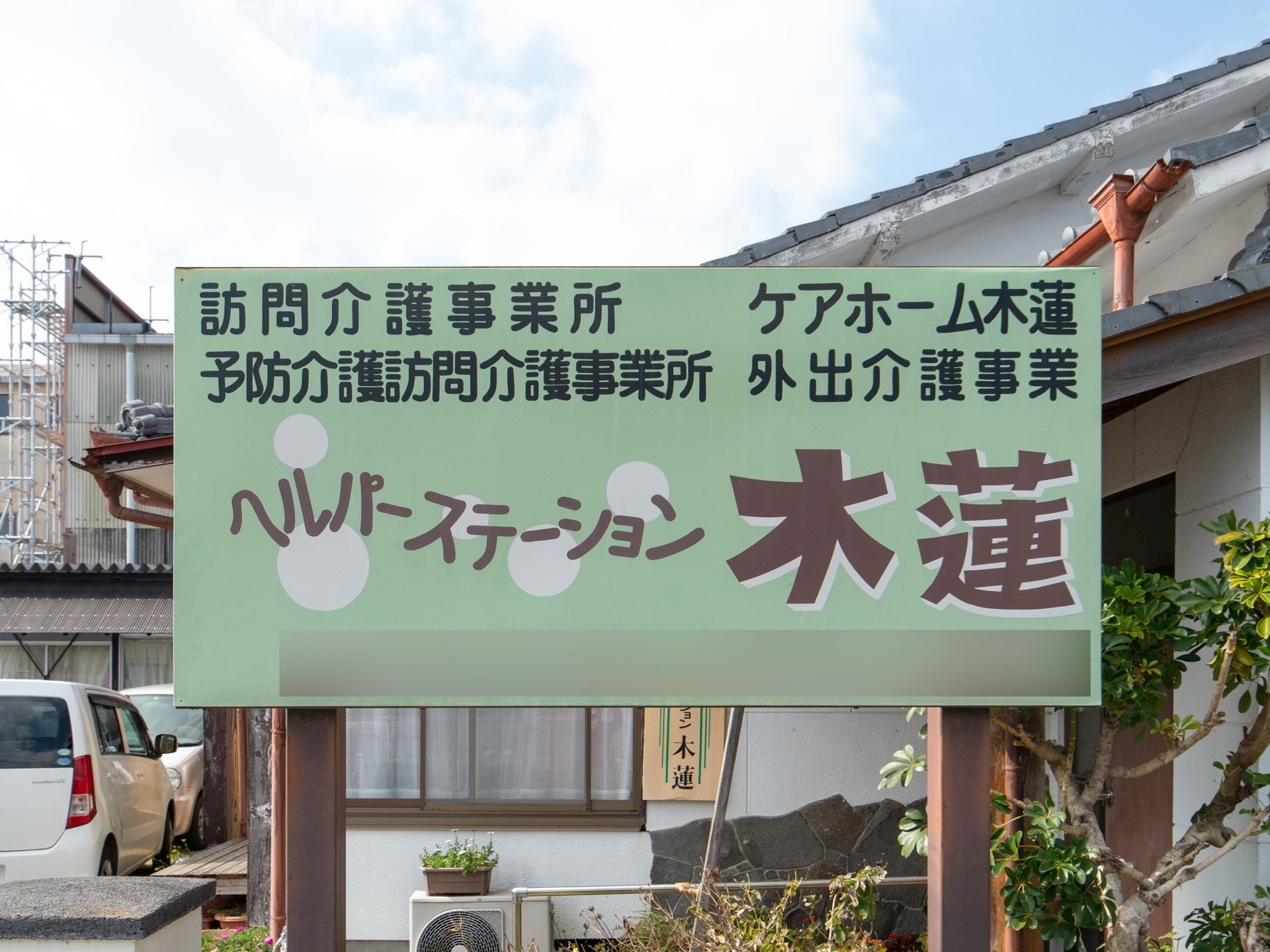 グリーン系の色調のプレートに、施設名称と事業種別がデザイン文字で明記され、施設建物の前に設置されている。