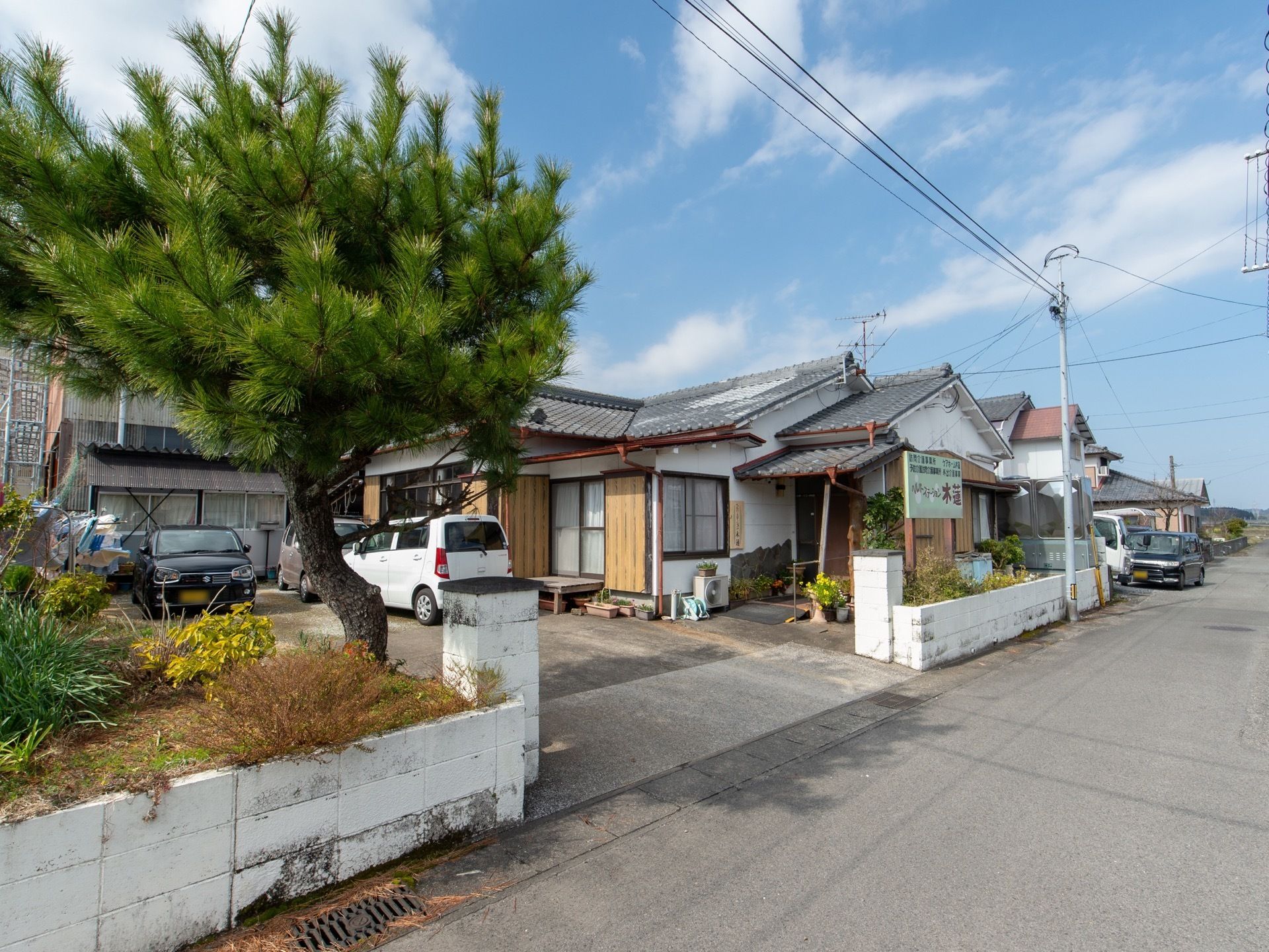晴れた日の木造一階建て施設の様子である。植栽を手前に駐車場、施設建物が見える。周囲には商業施設が点在している。