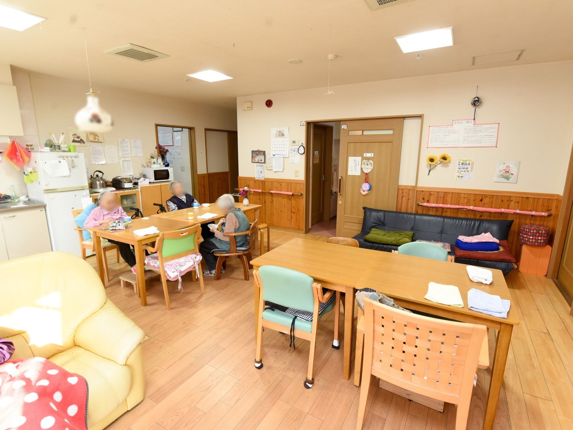 ４人で利用できるテーブルセットがあり、入居者様が座っている。奥に引き戸の扉があり、開けられている。手前に黄色のソファが１脚ある。