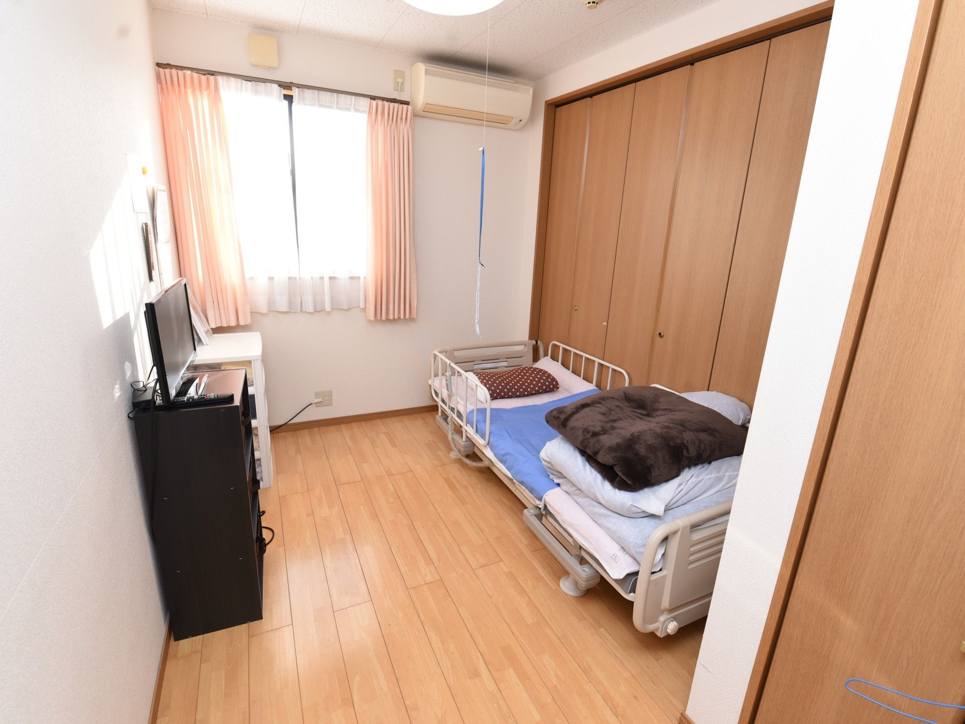 居室には大きな窓があり、ピンクのカーテンが下がっている。窓の横にエアコンが取りつけられている。壁側にチェストがある。