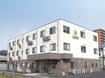 白とブラウンでまとめられた３階建ての建物。白い外壁には住宅の名前とロゴが描かれている。