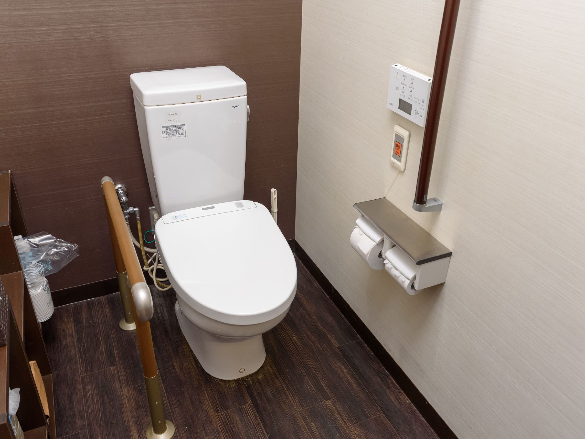 トイレの床と一部の壁は落ち着いた色合いの木目調である。白いトイレが設置されていて横に木製の手すりがある。