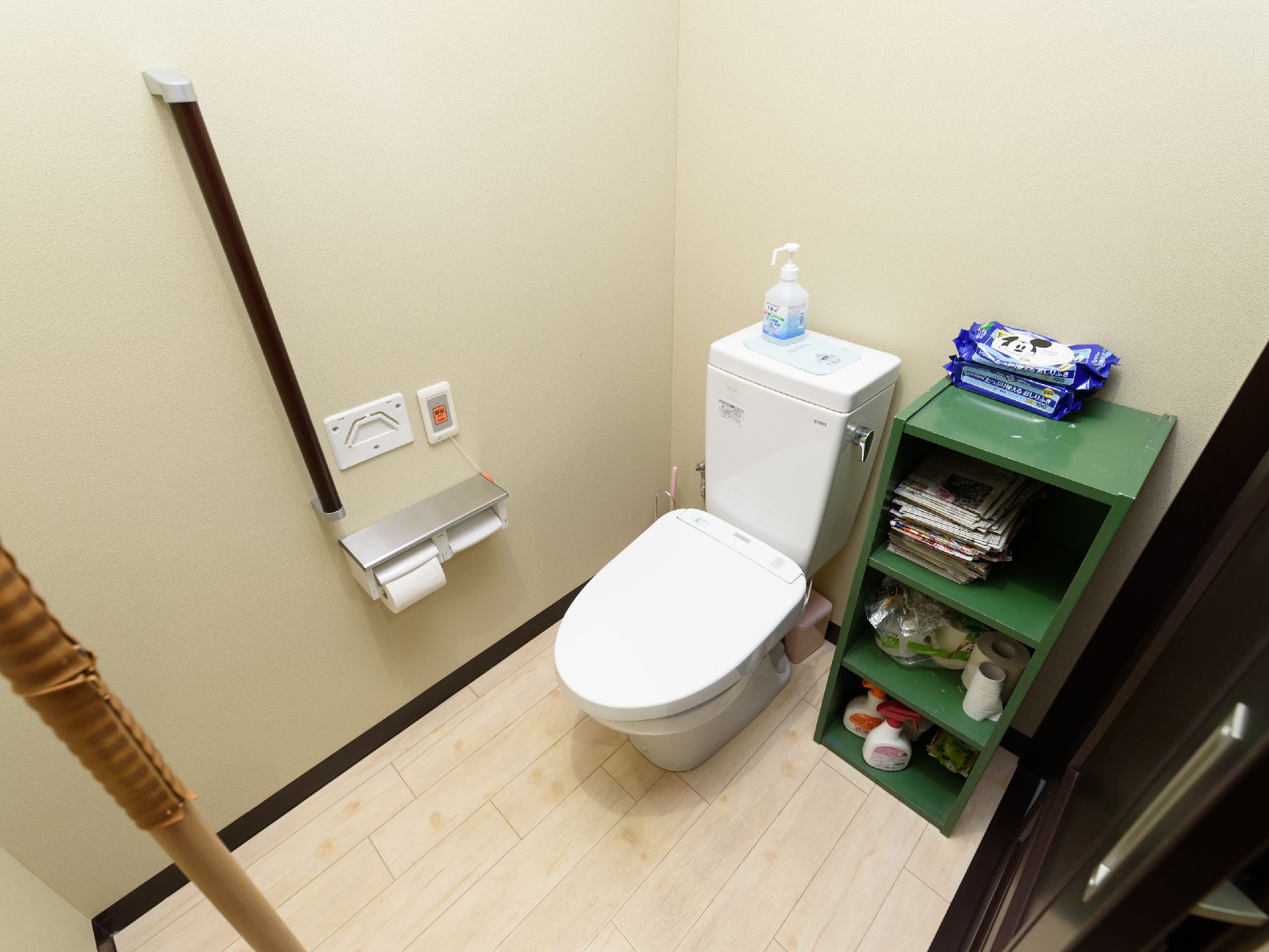 トイレの貯水タンクの上には青い液体が入ったボトルが置かれている。横に緑色の棚があり、洗剤やトイレットペーパーなどが入っている。