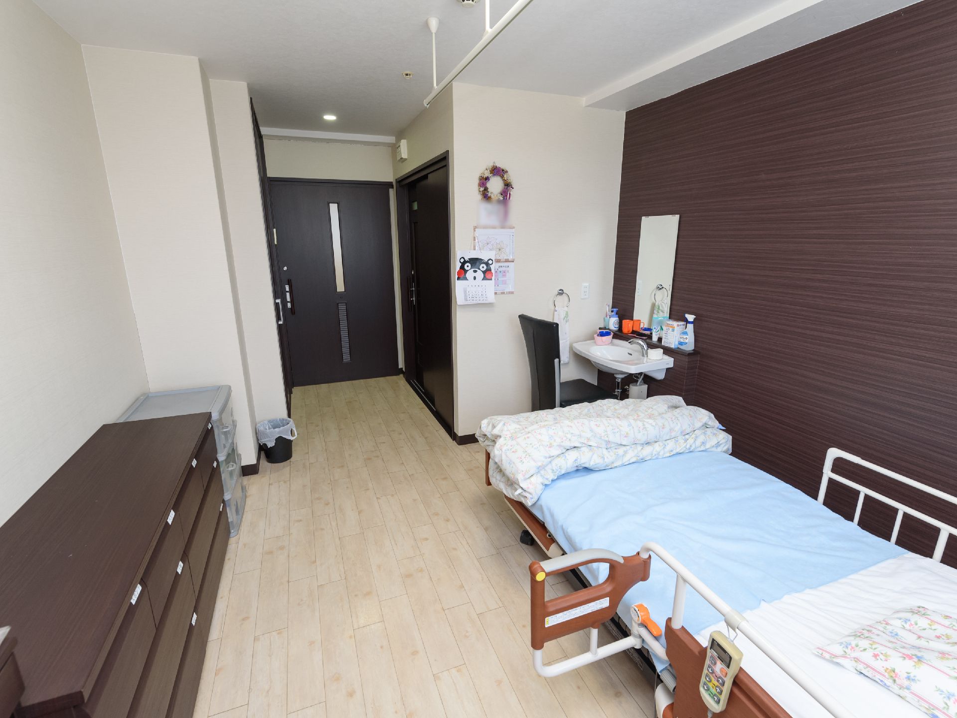 居室の一部の壁は濃い色合いの木目調となっている。介護用ベッドと収納家具、洗面台などが設置されている。