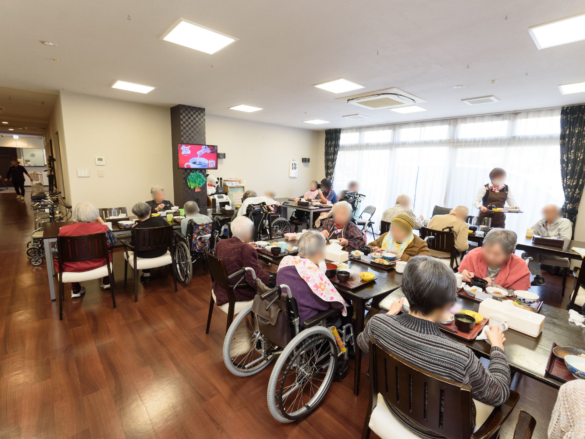 食堂で入居者様が大勢食事をしている様子である。それぞれが椅子に座ったり車椅子に座ったりしていて、テーブルに食事のトレーが乗っている。