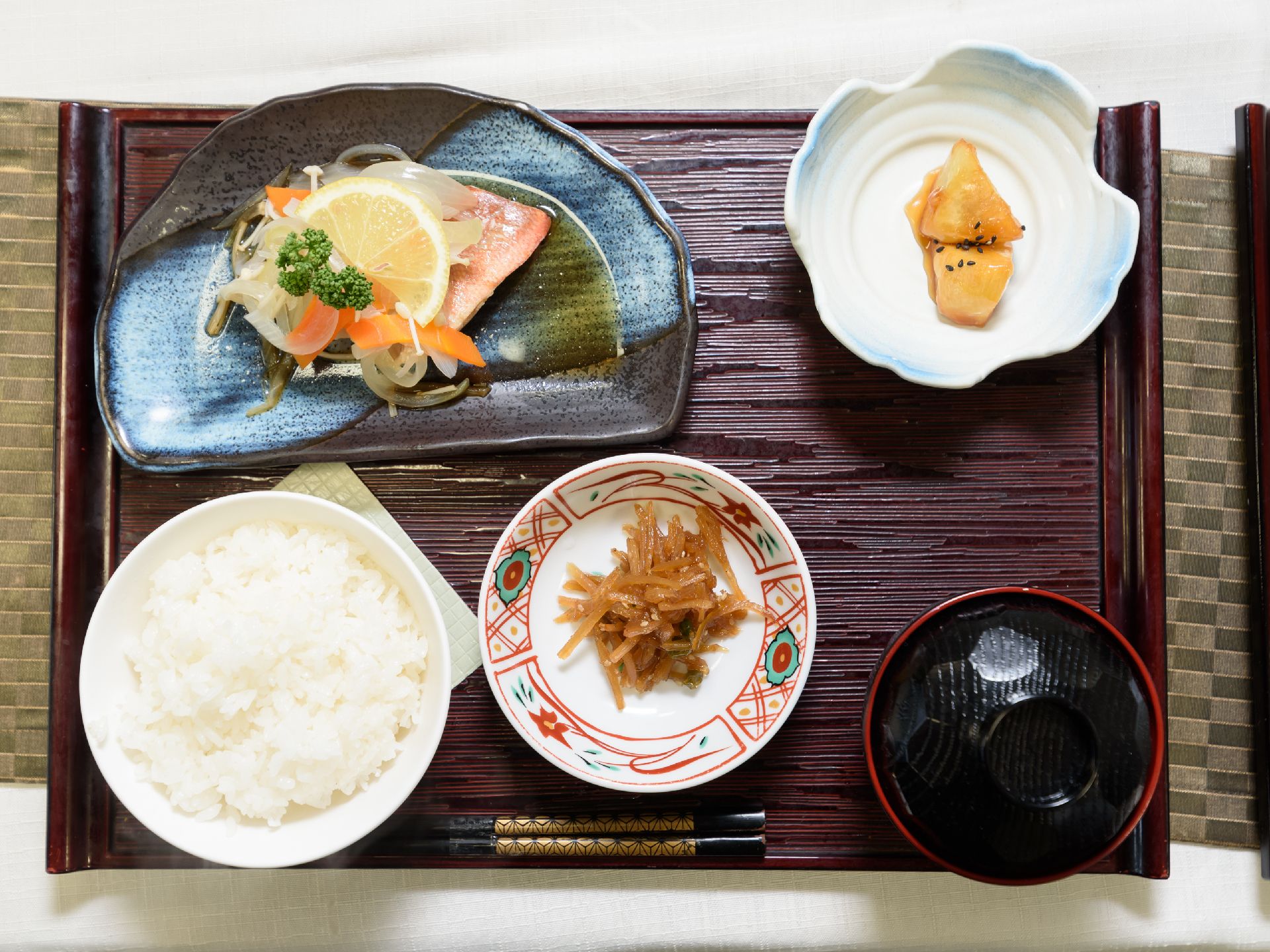 四角い木目調のトレーに食事が並べられている。手前に黒い箸があり、ご飯と味噌汁、おかずが３品乗っている。