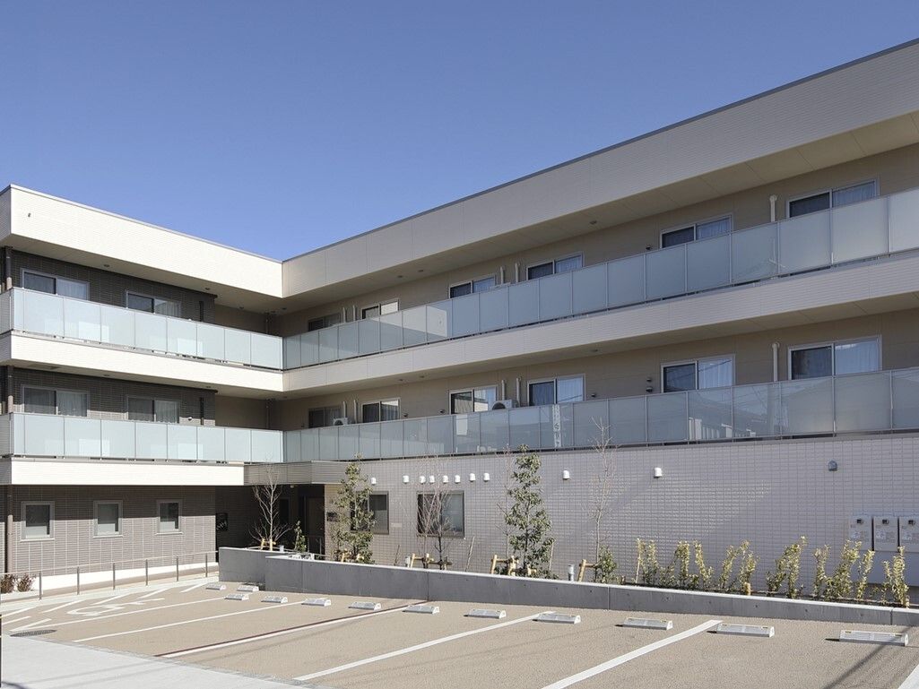 ベージュとダークブラウンの３階建てのエル型の外観である。建物の傍に植栽を取り入れ、横側に車を停めれるスペースを設けている。