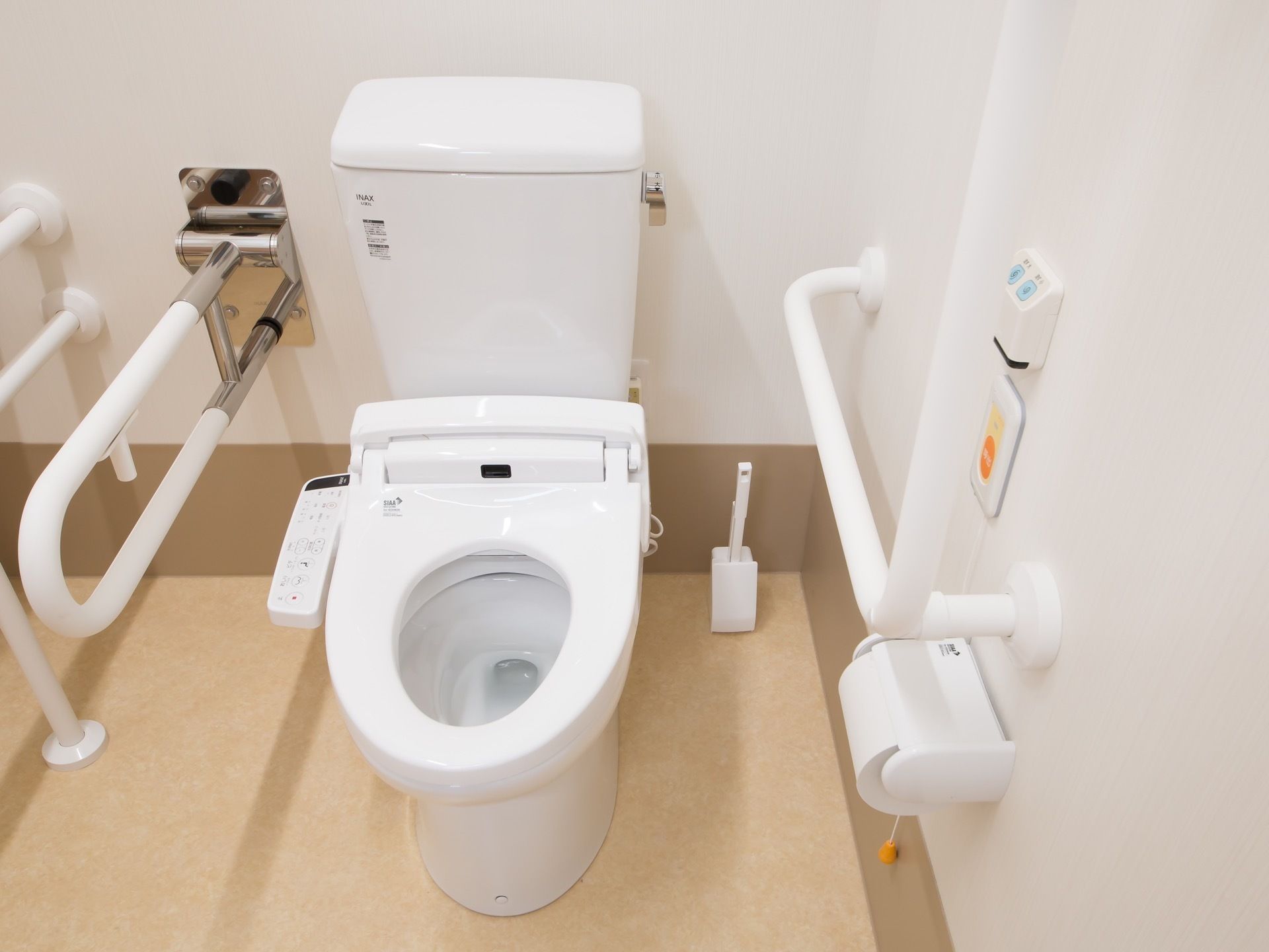 温水洗浄便座が設置された白を基調としたトイレである。操作パネルは便座の横にあり、その隣に可動式の手すりがある。