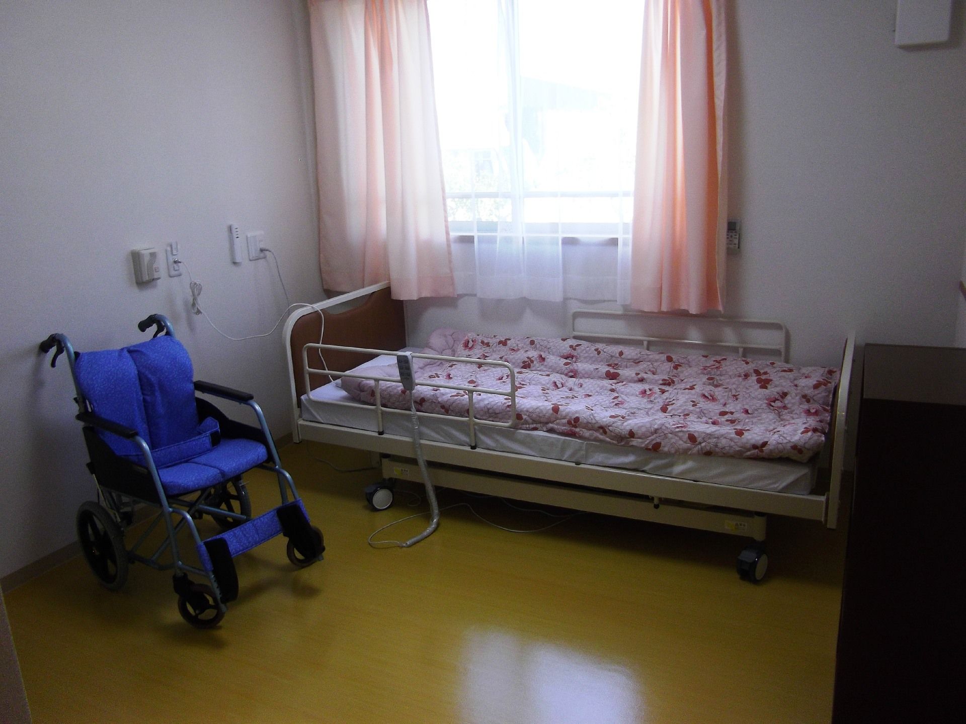 ピンクのカーテンが窓にかけられた居室には布団がセットされたベッドや収納家具、車いすが置かれている。