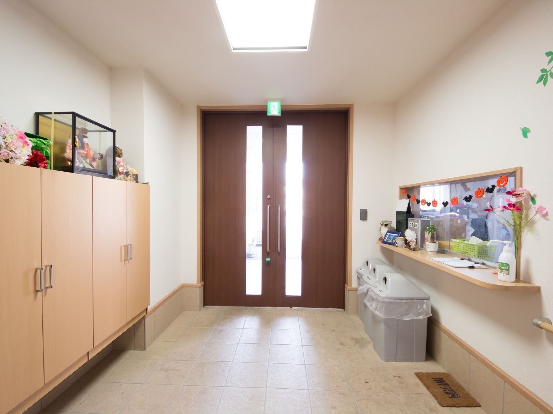 広い通路の片側に受付カウンターがあり、近くにごみ箱が3つ置かれている。反対側には扉付きの収納があり、正面のスライドドアの上には誘導灯がある。