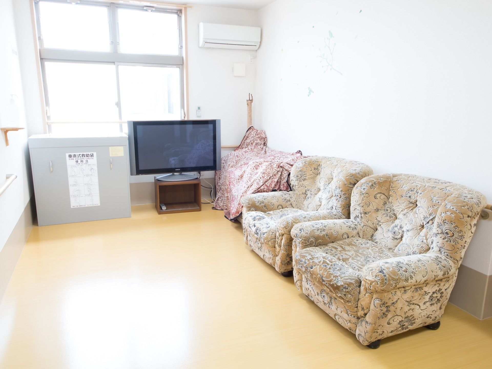 白い壁でベージュの床の部屋には窓から日光が射し込んでいる。エアコンとシングルソファーが2脚置いてあり、テレビもある。