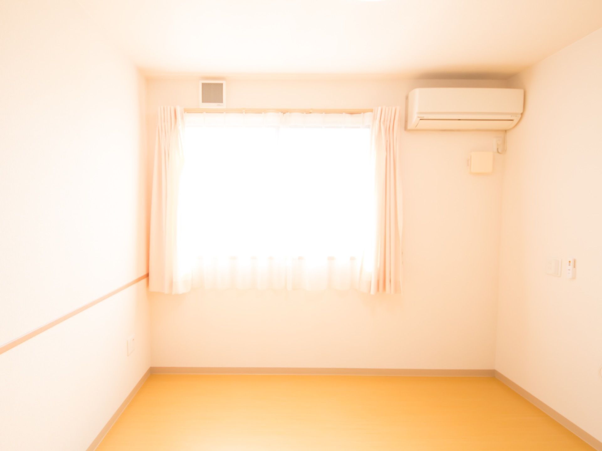 白い壁、ベージュのフローリングの床の部屋に窓から光が射し込み明るい。防炎カーテンとエアコンが設置されている。