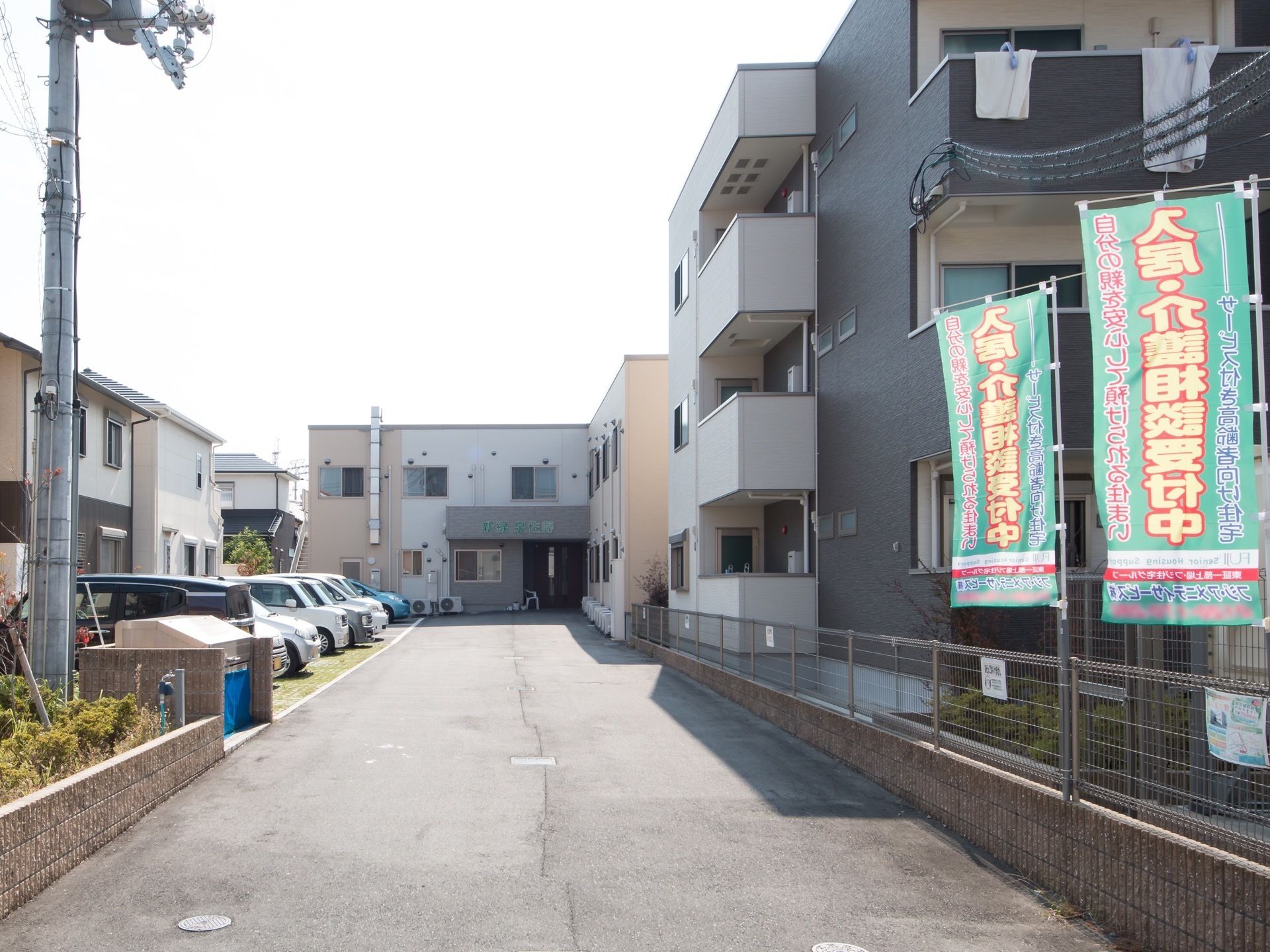 閑静な住宅街にある施設は2階建てで、手前の3階建の建物のフェンスに施設の入居者募集中の旗が立っている。道の端が駐車場になっている。