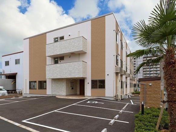 3階建てのすっきりとした建物は白にブラウンのアクセントがある。ヤシの木などが植えられた駐車スペース。