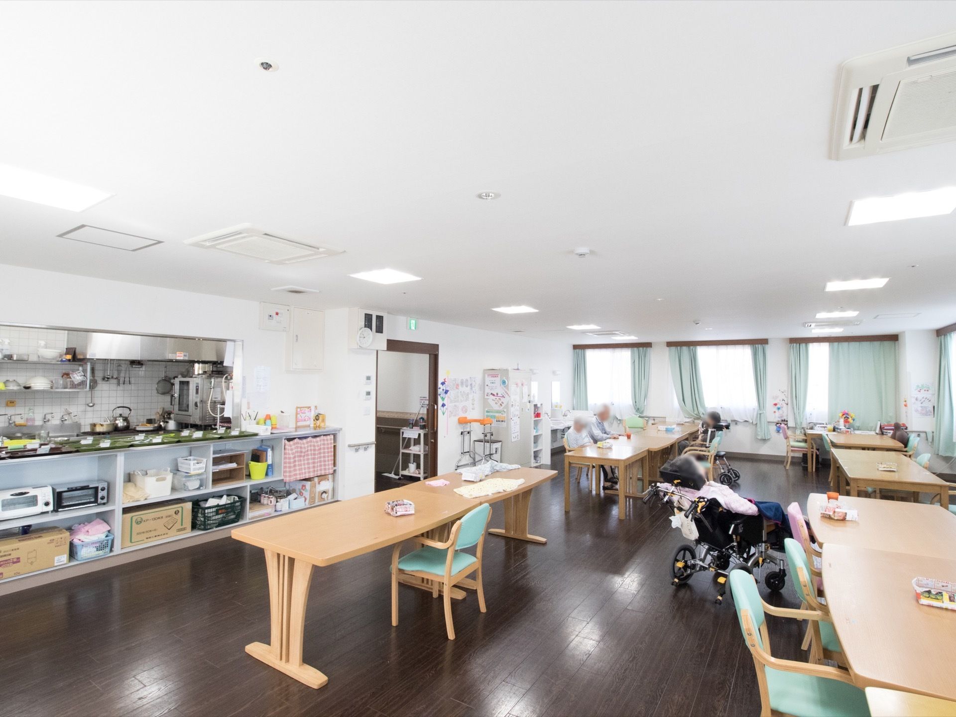横長のテーブルが複数置かれており、その周りに椅子が並べられている。入居者様が椅子に座り過ごされている。