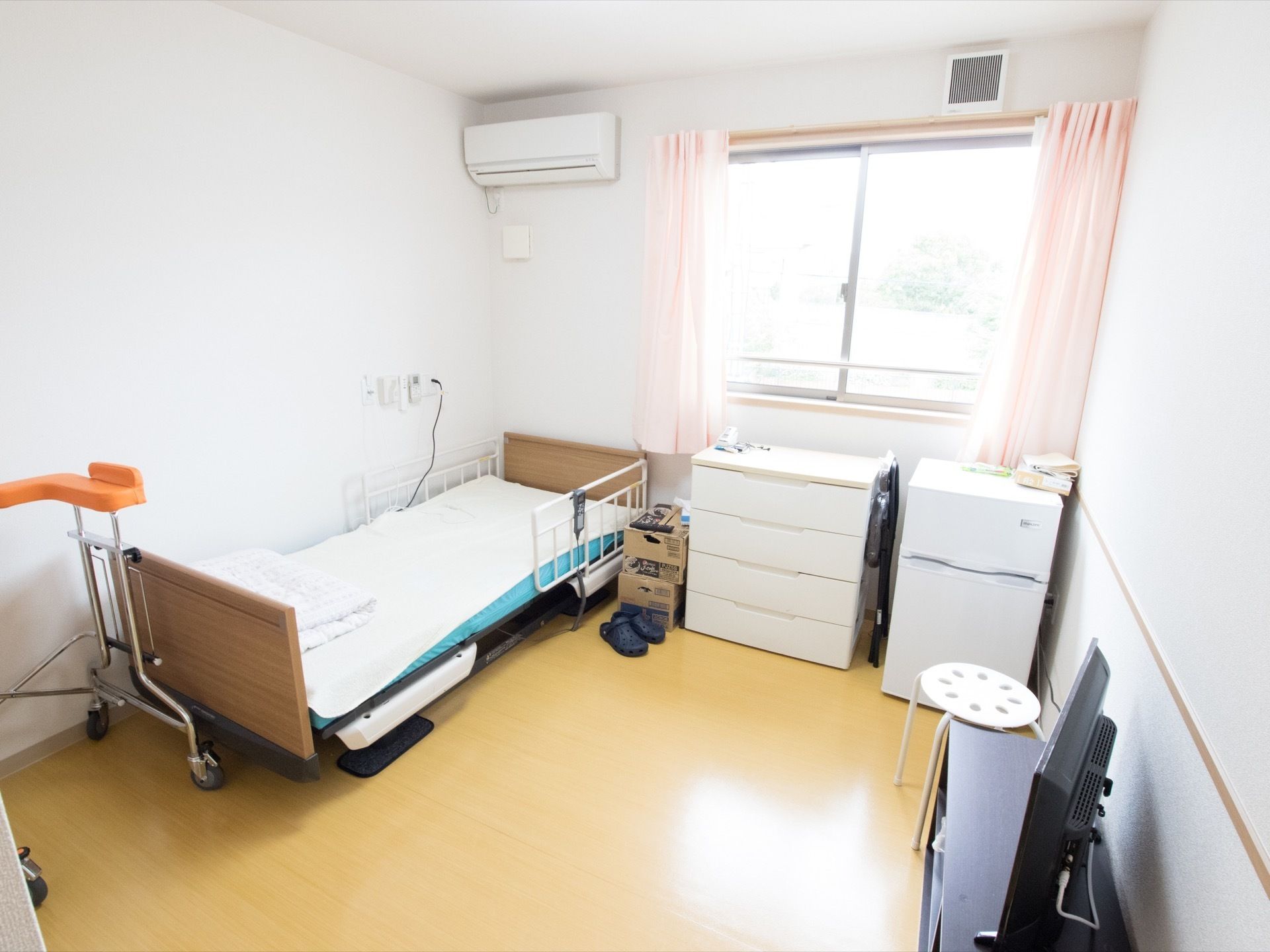 居室には壁際にベッドがあり、ピンクのカーテンがかかった窓の下にチェストと冷蔵庫が置かれている。エアコンが完備されている。