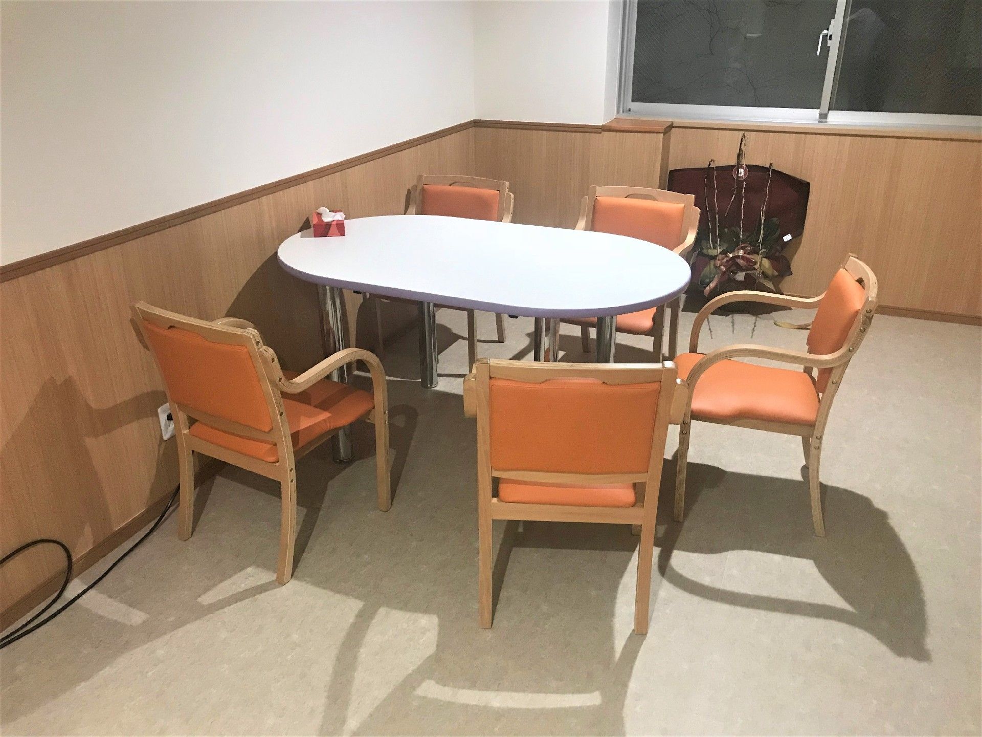 白い机にオレンジ色の椅子が5脚置かれている。机は円形で、机を囲むように椅子が置かれている。