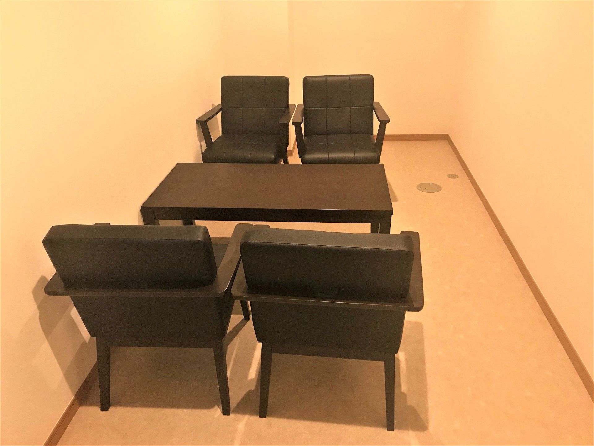 白い壁と床の部屋にこげ茶色の机と椅子が置かれている。暖色系の照明が使われており、温かみのある部屋になっている。