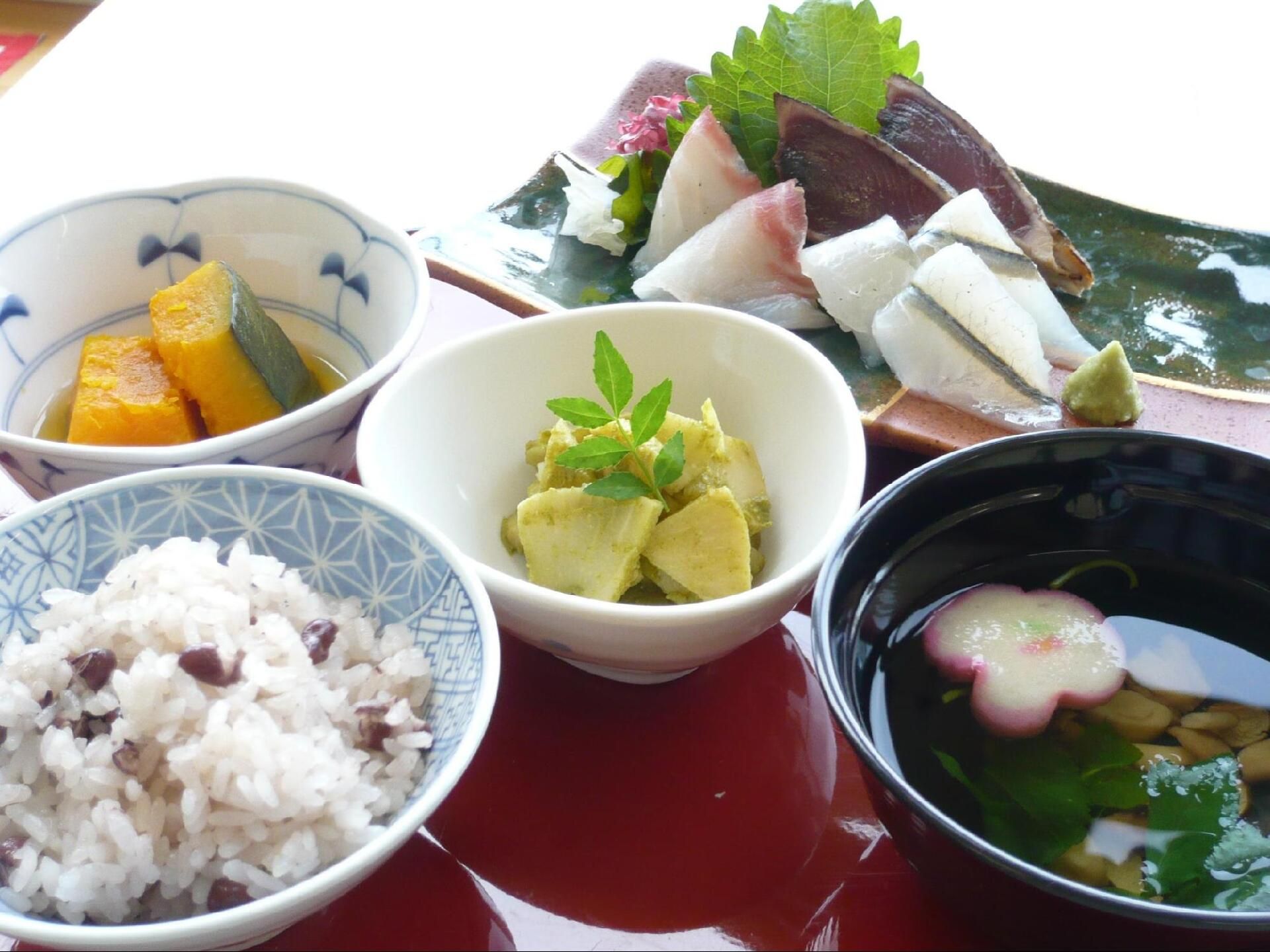 ３種類の刺身の盛り合わせと赤飯、かぼちゃの煮物、タケノコの木の芽和えなどが並べられている。