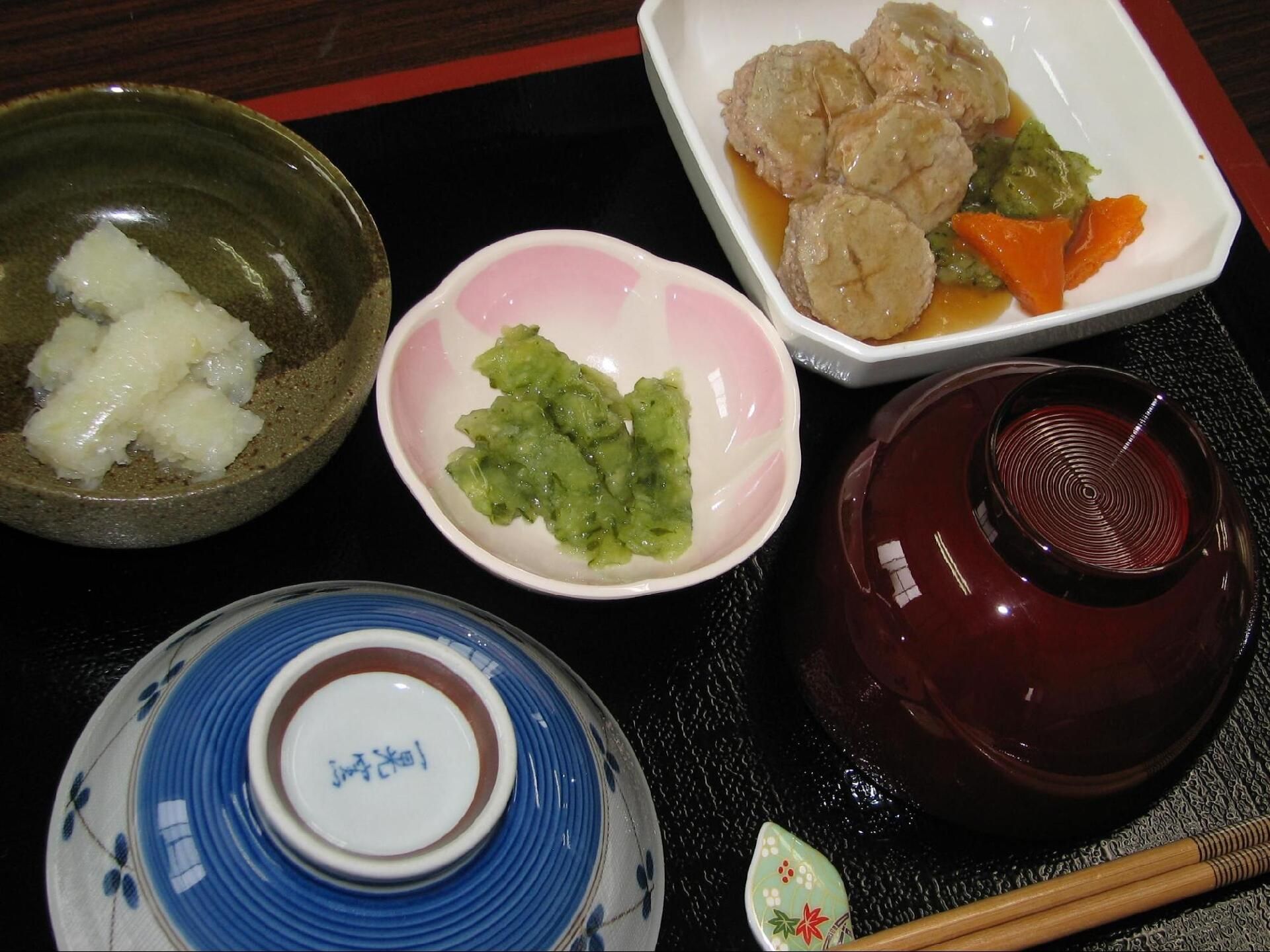 汁物の椀が右手にあり、茶碗が左手にあり伏せられている。中央には緑の食材、左上には白い棒状のもの、右上には煮物のソフト食がある。