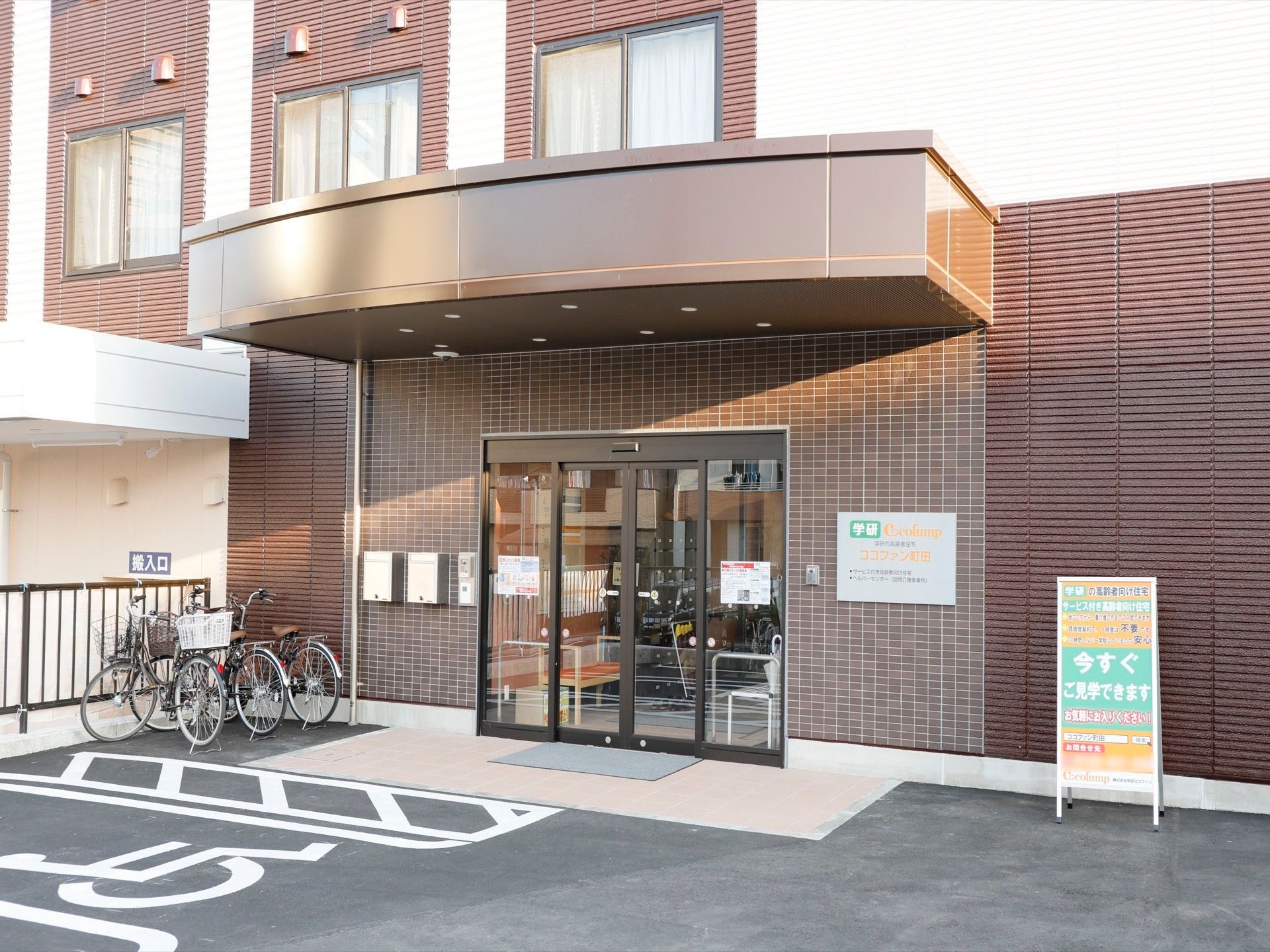 ダークブラウンとベージュのツートンカラーの外壁が落ち着いた雰囲気の玄関である。入り口の傍に見学案内の看板が置かれている。