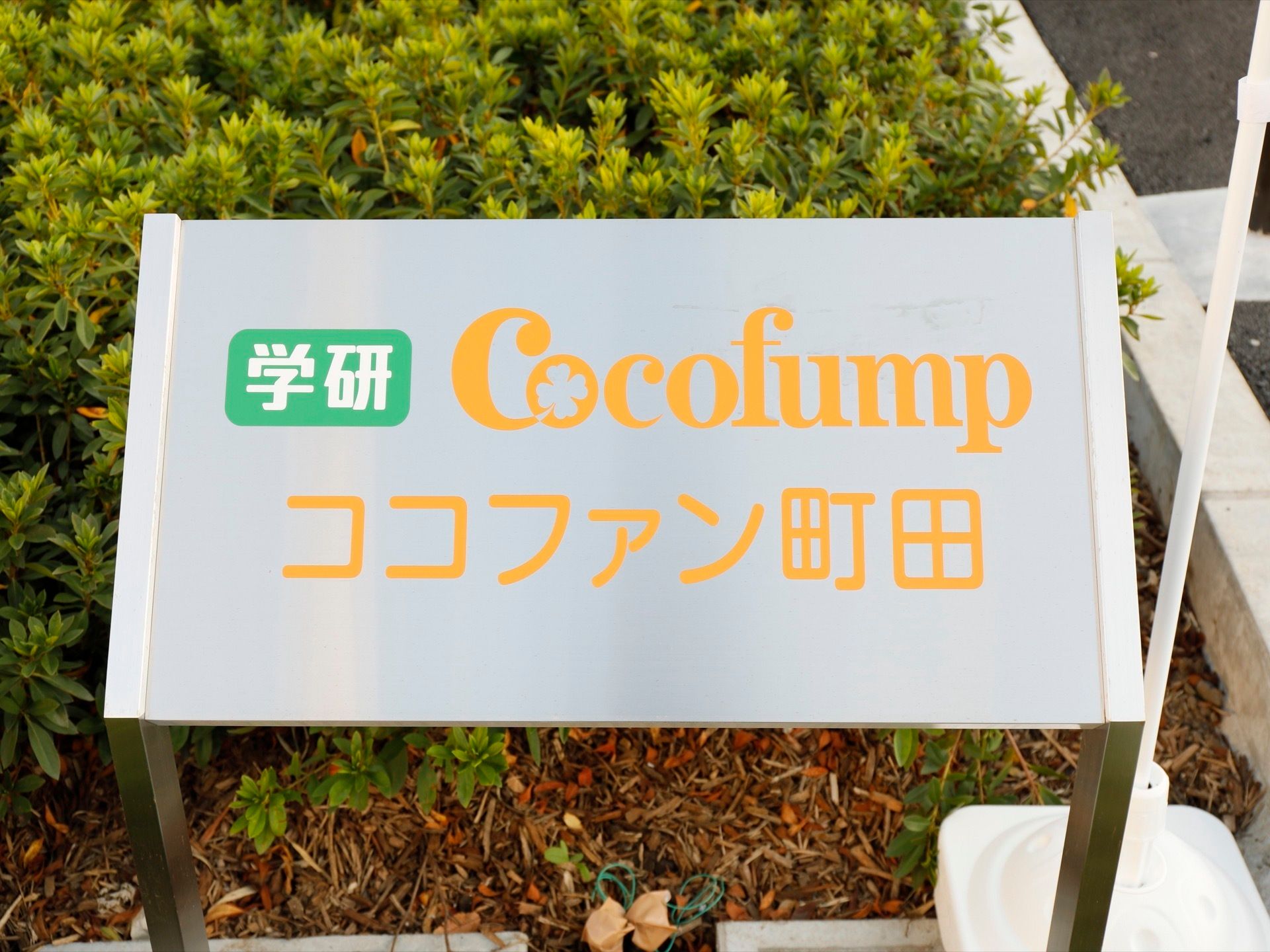 白いレンガで囲われたスペースに、丈の低い植物が植えられている。オレンジで施設名の書かれた銀色のプレートが設置されている。