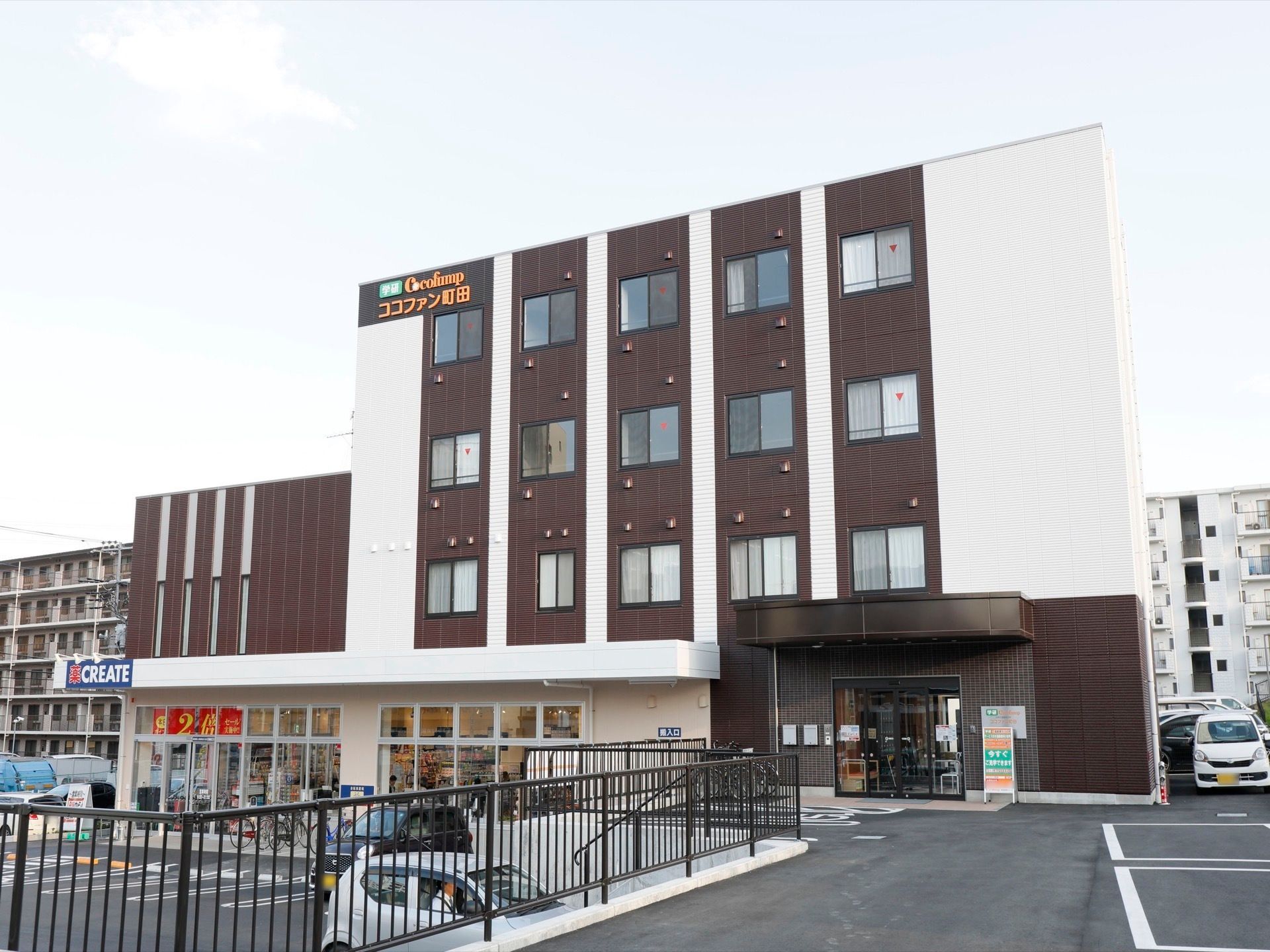 ダークブラウンの外壁に施設の看板が掛けられている。玄関を出てすぐに駐車場が設けられており、隣にはドラッグストアがある。