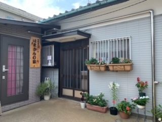 施設の隣には住宅が建てられており、間にはブロック塀が建てられている。玄関ドアの両脇にはプランターなどに植えられた植物が飾られている。