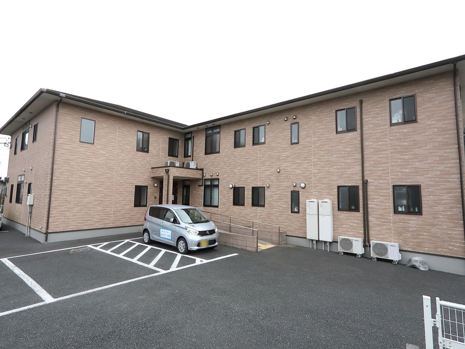 直角に２方向に分かれたお部屋を持つ建物。駐車場前にスロープがある。外壁は茶系で、サッシなどの枠は黒い素材を用いている。