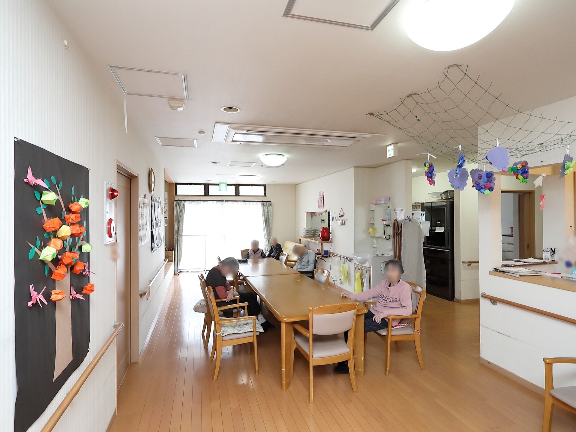 ダイニングテーブルと椅子がセットで二組並べられており、入居者様が数名椅子に座っている。壁には折り紙で作られた飾りが掛けられている。