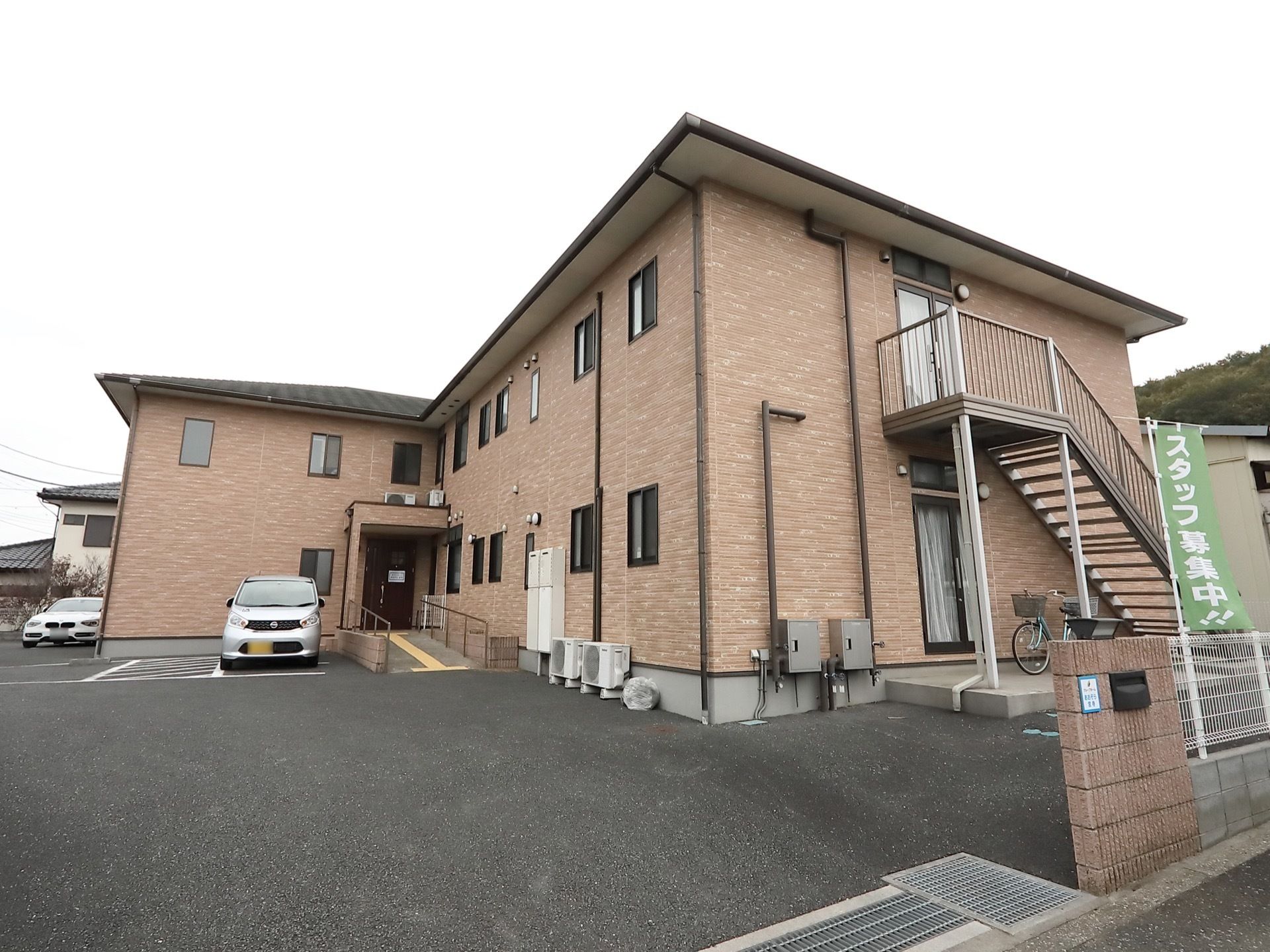 二階建ての建物が見える。非常階段が用意されている。施設入り口まではスロープになっている。車が数台駐車している。