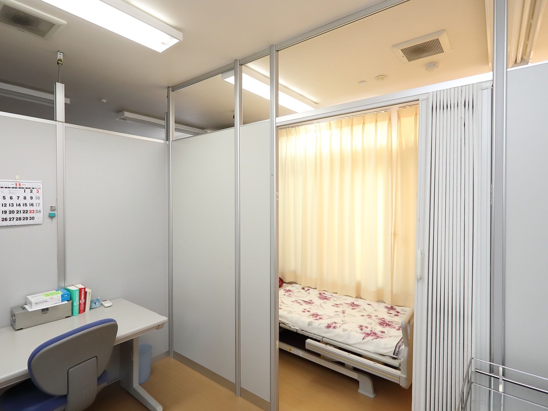 パネルに仕切られた部屋に机と椅子が置かれている。アコーディオンカーテンに仕切られた先の小部屋にはベッドが置かれている。