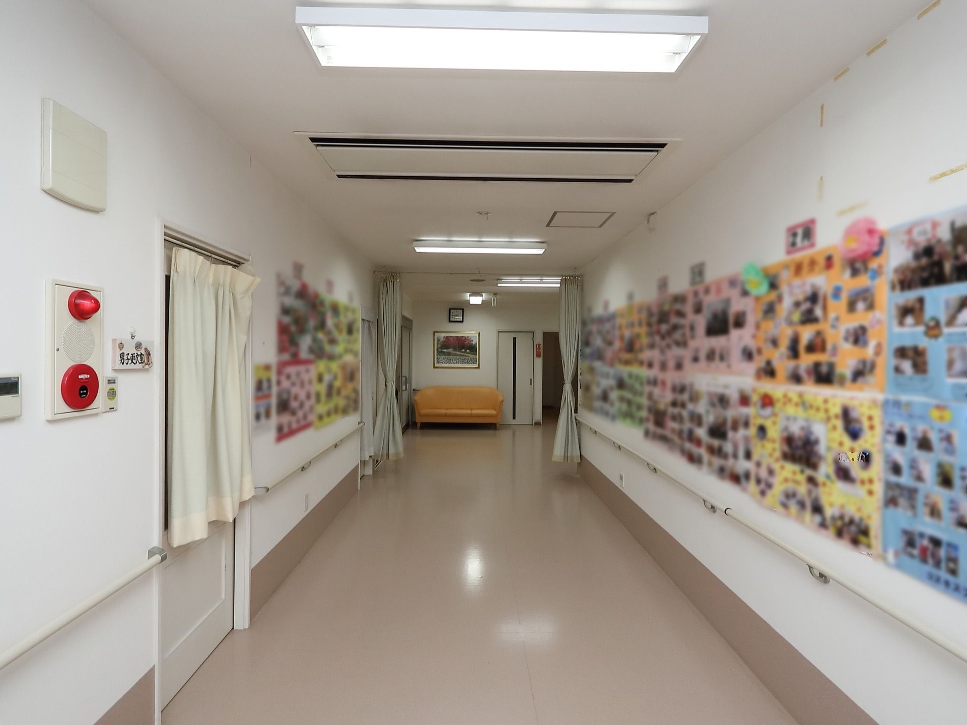 壁に色とりどりの台紙が貼られ、入居者様の写真が飾られている広い廊下。突き当りにはオレンジのソファが置かれている。